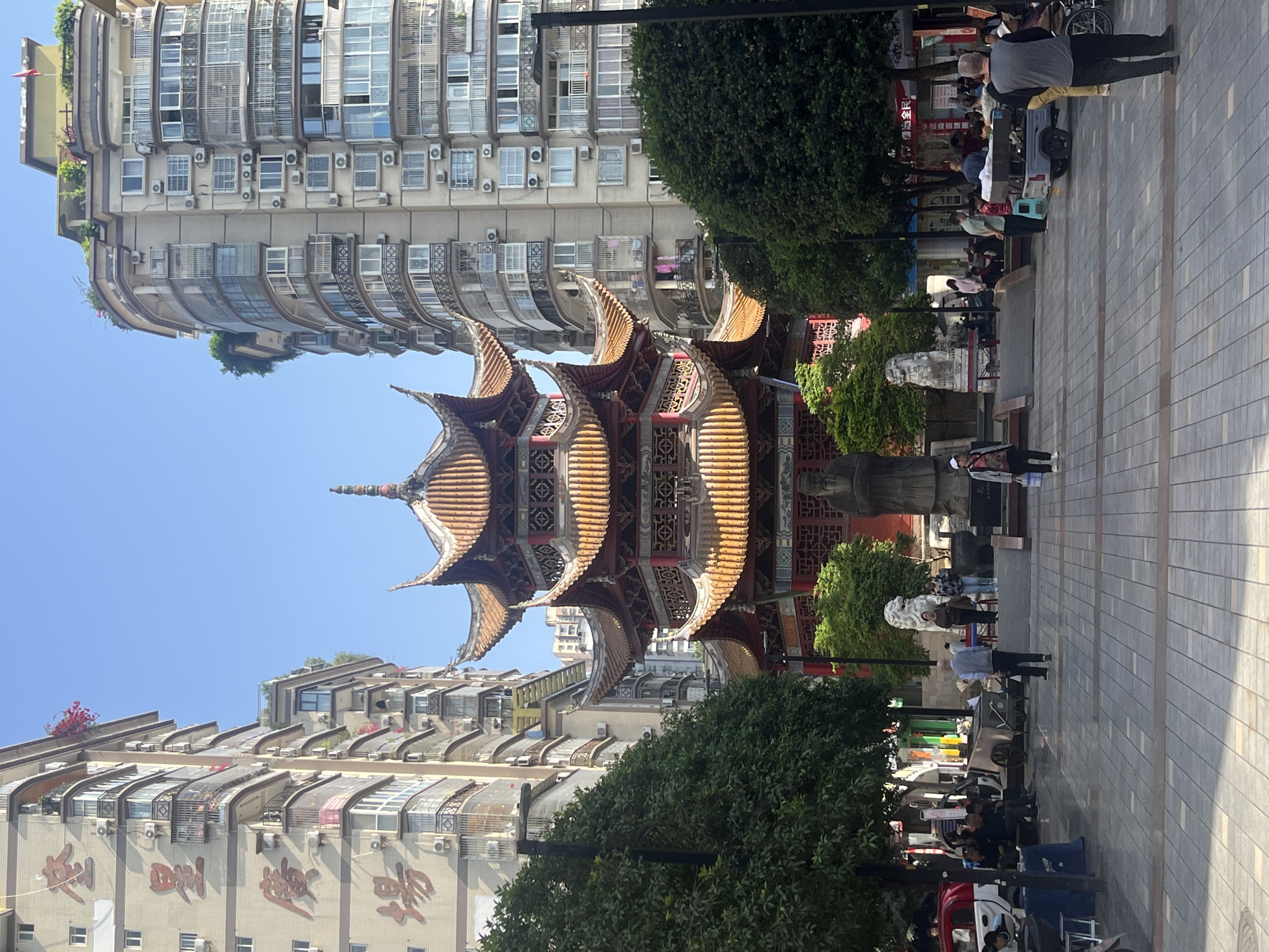 This image captures a vibrant urban scene in Jiangjin, China, on a bright, sunny day. The central focus is a magnificent, multi-tiered traditional Chinese pagoda-style building, characterized by its ornate golden-brown tiled roofs, intricate carvings, and striking architectural details. It stands as a cultural landmark amidst contemporary urban development.
Flanking the traditional pagoda are modern, multi-story apartment buildings. The building on the left features a distinctive design with numerous curved balconies, large windows, and an abundance of air conditioning units. On its facade, prominent red Chinese characters are visible, which appear to read "江津商业街" (Jiangjin Commercial Street), indicating the area's commercial identity.
In the foreground and extending to the right, a paved pedestrian square is bustling with activity. Various people are seen walking, standing, and sitting, engaged in their daily routines. Some individuals are pushing carts or strollers, suggesting a mix of locals and visitors. The square is dotted with green trees and bushes, providing shade and natural elements to the otherwise built environment. The clear blue sky overhead confirms fair weather conditions. The scene effectively portrays a lively public space where traditional architecture coexists with modern urban living.