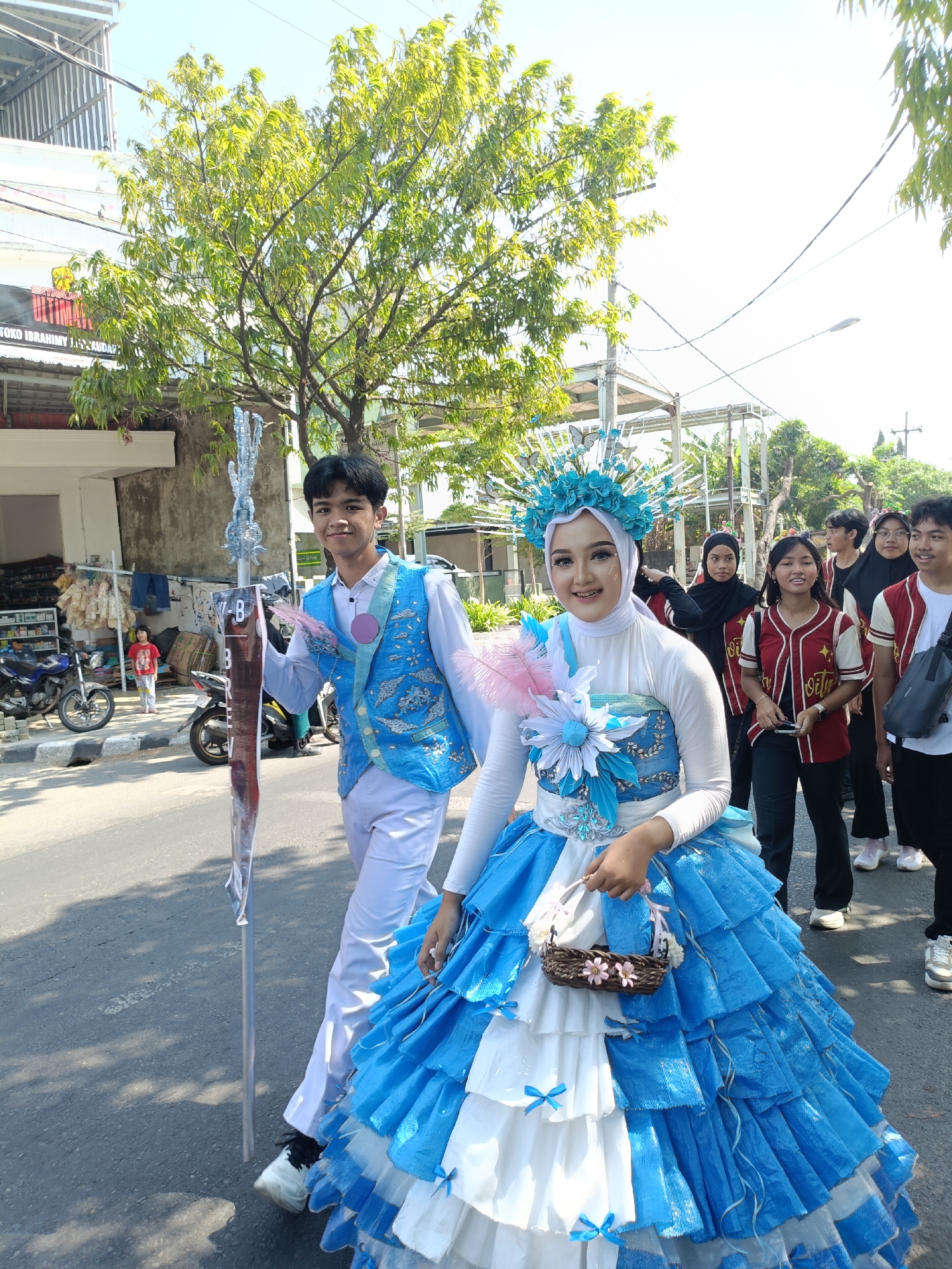 Two young individuals, dressed in traditional performance attire, are participating in a street parade in Kediri City, Indonesia. The female on the right is wearing an elaborate blue and white ruffled gown and an ornate headdress adorned with blue flowers and butterflies. She holds a small woven basket with pink flowers. The male on the left is wearing a white shirt, a blue vest with intricate embroidery, and white pants. He is holding a tall staff decorated with a crystalline finial. Behind them, a group of young people in casual clothing, some wearing maroon and white vests, follow along the street. The background features a building with a red sign, trees, power lines, and parked motorcycles, indicating an urban setting. The ground is asphalt, suggesting a road. The lighting suggests it is daytime, with bright sunlight casting shadows.