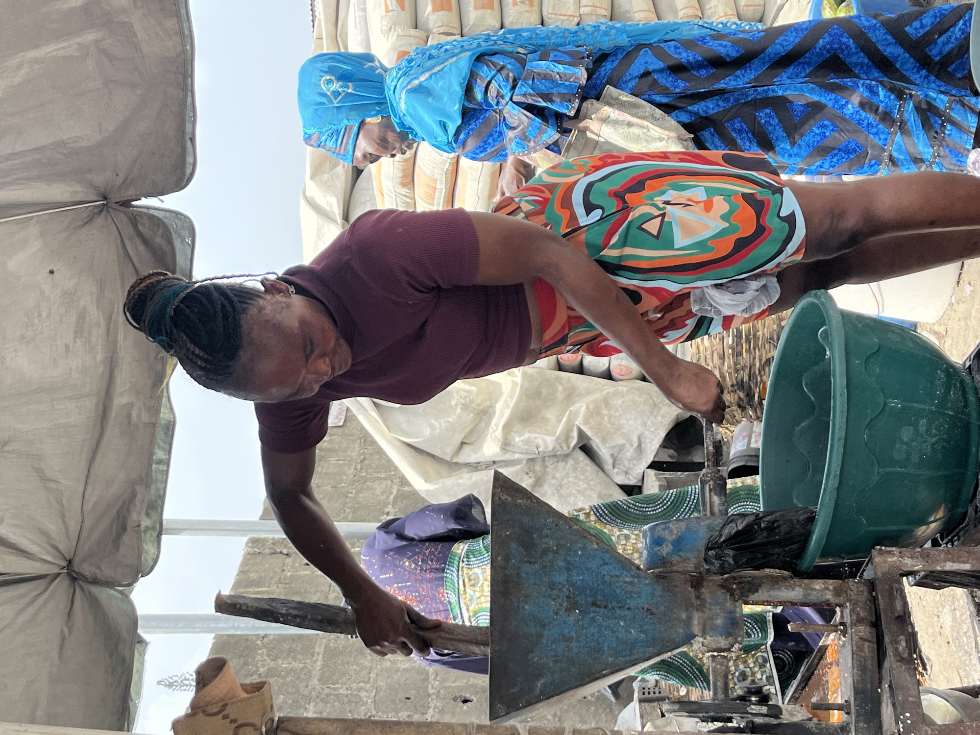 This image, rotated 90 degrees clockwise, captures an outdoor or semi-outdoor scene featuring two women engaged in a processing activity.
In the foreground, an adult woman with dark skin and braided hair styled in a bun is bent over, actively operating a machine. She wears a dark red short-sleeved top and vibrant, multi-colored patterned shorts or a short skirt with geometric designs in orange, green, blue, black, white, and brown. She holds a wooden stick, appearing to feed material into or manipulate the contents of a blue and metal machine, which resembles a grinder or processor.
Behind and to the right of her stands another adult woman with dark skin. She wears a light blue head covering, possibly a hijab or scarf, adorned with heart-like embroidered motifs, and a long blue and black patterned garment. She is looking down, seemingly observing the activity.
Adjacent to the machine, a large, dark green plastic basin or bucket contains a black plastic bag, likely positioned to collect processed goods. In the background, numerous white or light-colored sacks are stacked, possibly containing grains or other bulk products, though any text on them is illegible. A light-colored tarpaulin or fabric forms a part of a shelter or awning on the left side of the frame. A rough brick wall is visible in the background. The bright, overcast sky indicates it is daytime. The scene depicts a focused and industrious activity in a local setting, consistent with its location in Ado, Nigeria.