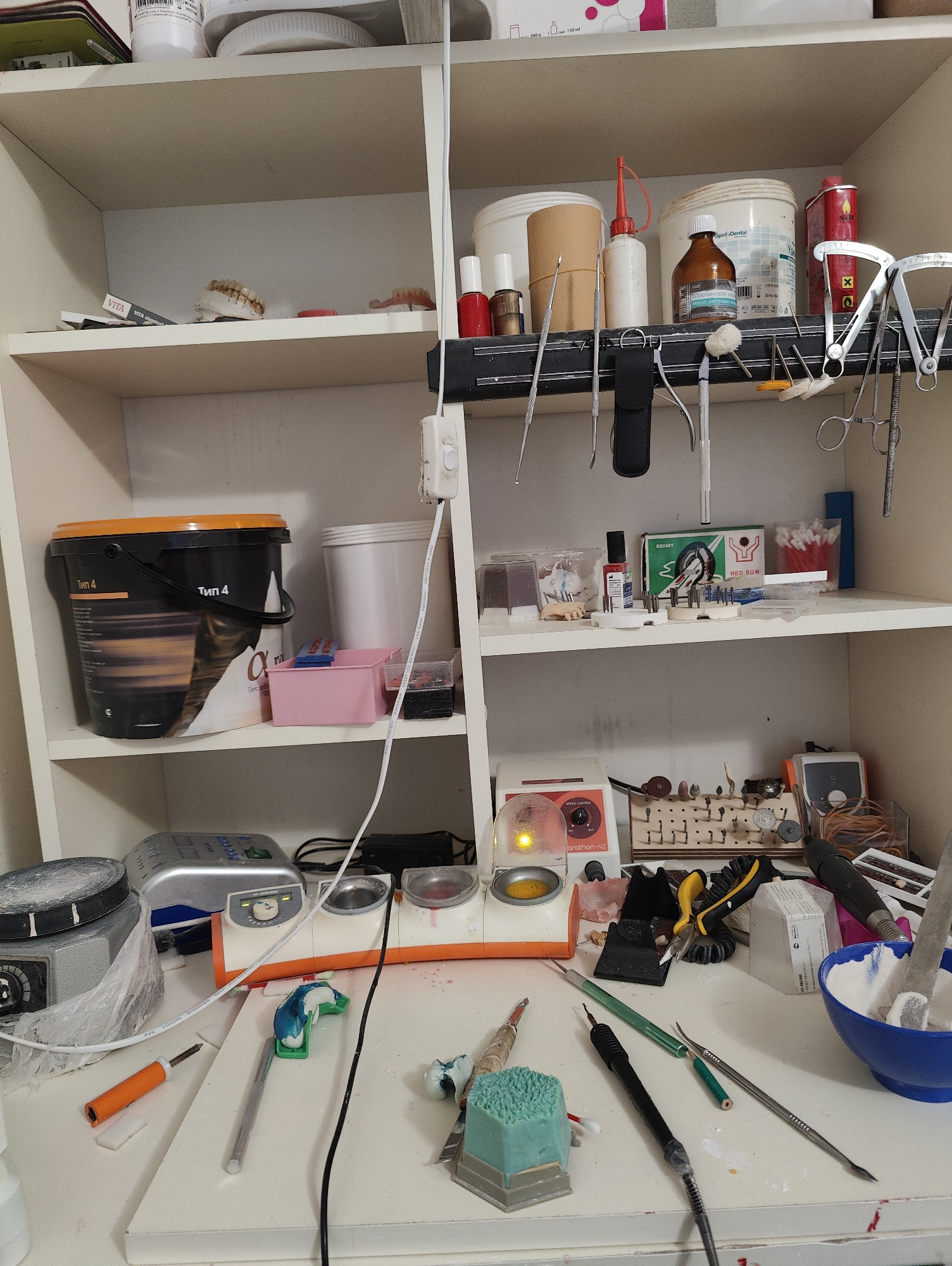 The image depicts a cluttered workbench in what appears to be a dental laboratory. The workbench is laden with various tools, equipment, and materials, suggesting ongoing work. Items visible include dental instruments, polishing tools, a dental casting machine, various containers, and what seem to be dental prosthetics. The overall impression is one of a functional workspace, possibly in Nuradilovo, Russia, though there is no direct evidence to confirm this location. The lighting suggests an indoor setting.