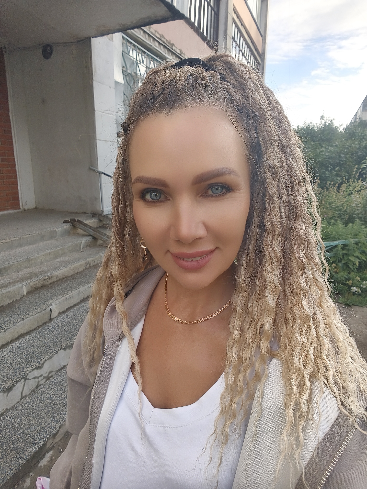 A woman with long, curly blonde hair stands outside a building. She is wearing a grey zip-up jacket over a white t-shirt and a gold chain necklace. She has blue eyes and is smiling. In the background, there are concrete stairs and a building with windows. There are also trees and a cloudy sky.