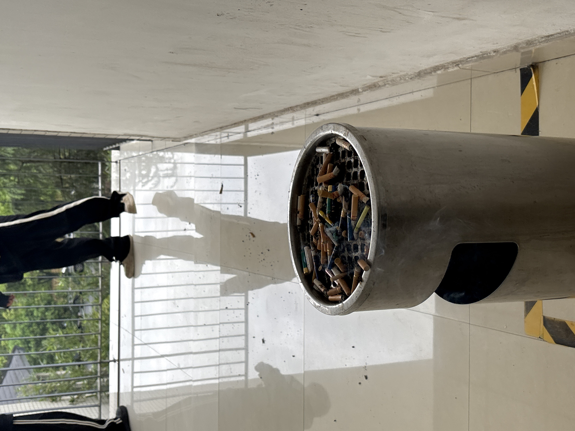 The scene is an outdoor smoking area, presumably in Jiangjin, China. A cylindrical metal ashtray mounted to a wall is filled with cigarette butts of varying colors and brands. A person is standing near a fenced-in balcony. The area is a tiled floor. A yellow and black stripe is seen next to the cylinder. The picture appears to have been taken from above.