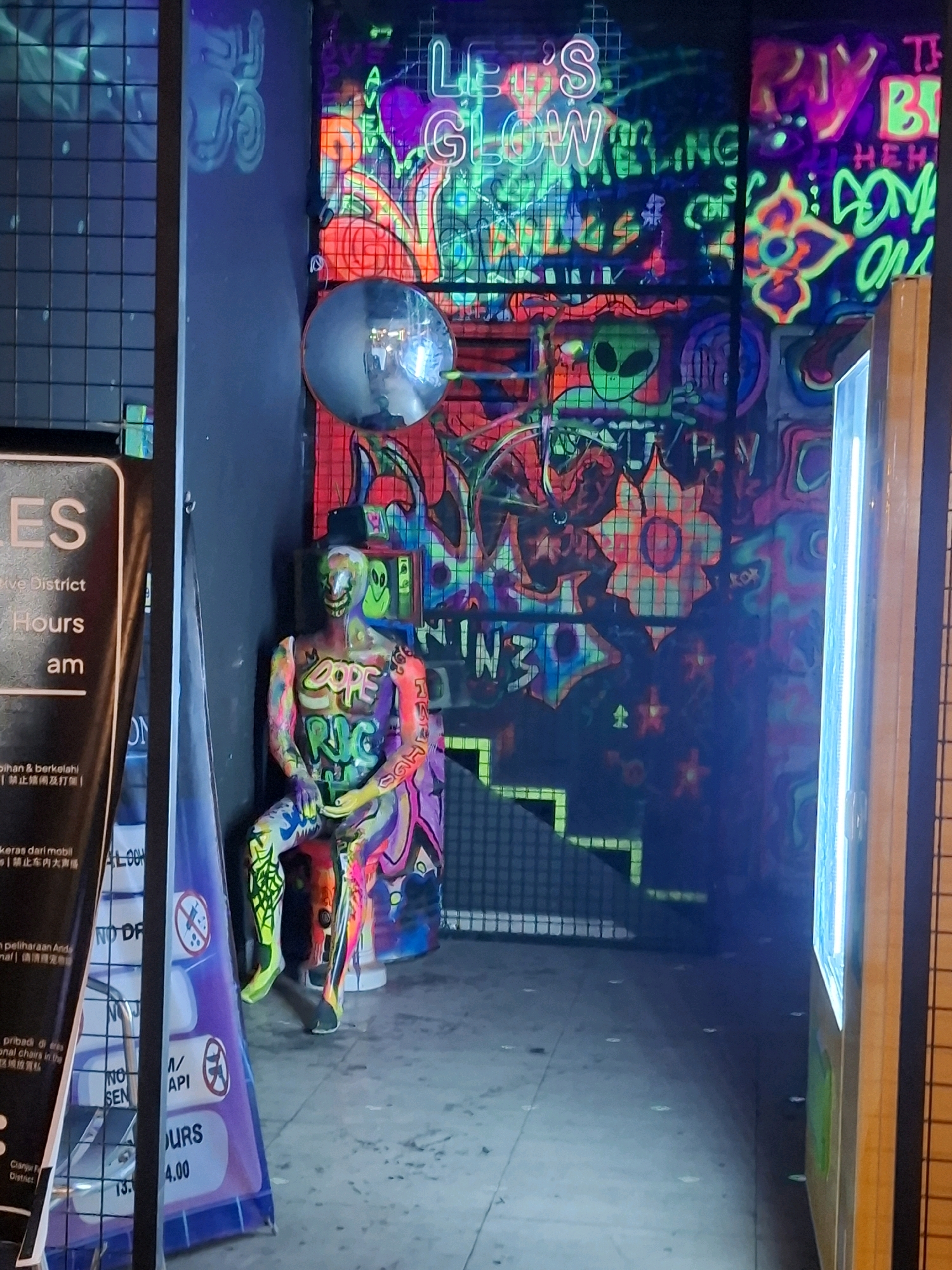 The image depicts a dimly lit interior space adorned with vibrant neon graffiti and artwork. In the foreground, a colorful, painted mannequin is seated on a small platform. The mannequin is covered in a chaotic pattern of bright, fluorescent colors and various symbols, including the word "DOPE" emblazoned across its torso. To the left of the mannequin, a black sign with white text is partially visible, displaying information about "District Hours" and "am," suggesting it might be a signage for a business or establishment. Further to the left, another purple sign with white icons and text, including "NO SMOKING" and "NO DRINKING," is also visible. The background features a wall covered in elaborate neon graffiti with various designs, including an alien figure, flowers, and text that appears to read "LET'S GLOW." A reflective sphere, possibly a mirror or decorative element, is mounted on the wall above the mannequin. The overall scene has a dark, psychedelic, and artistic ambiance, likely part of a nightlife venue or a themed exhibition space in the city of Cianjur, Indonesia.
