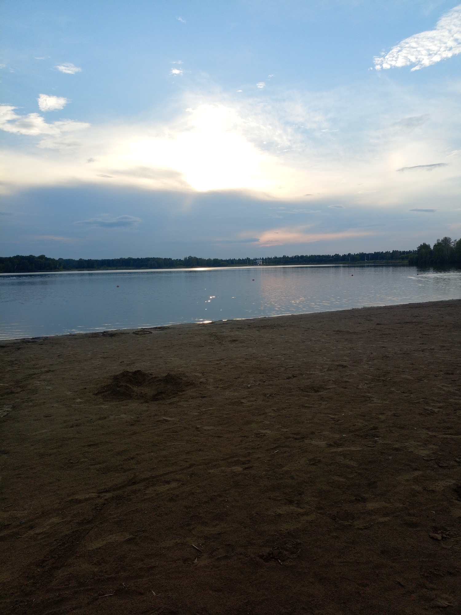 The image shows a wide, calm body of water, likely a lake, bordering a sandy beach. The sky above is a mix of blue with scattered white clouds, and the sun is partially obscured, suggesting it is either early morning or late evening. The water reflects the sky and surrounding trees, which form a dark green line along the distant shore. The beach in the foreground is sandy and appears to be a public area, possibly a designated swimming or recreational spot. There are no people visible in the immediate foreground, but a few distant buoys or markers are visible in the water. The overall scene is peaceful and natural, evoking a sense of tranquility. This location is likely the beach area of Lake Serebryanoye (Silver Lake) in Zheleznogorsk, Russia, a popular recreational spot within the city.