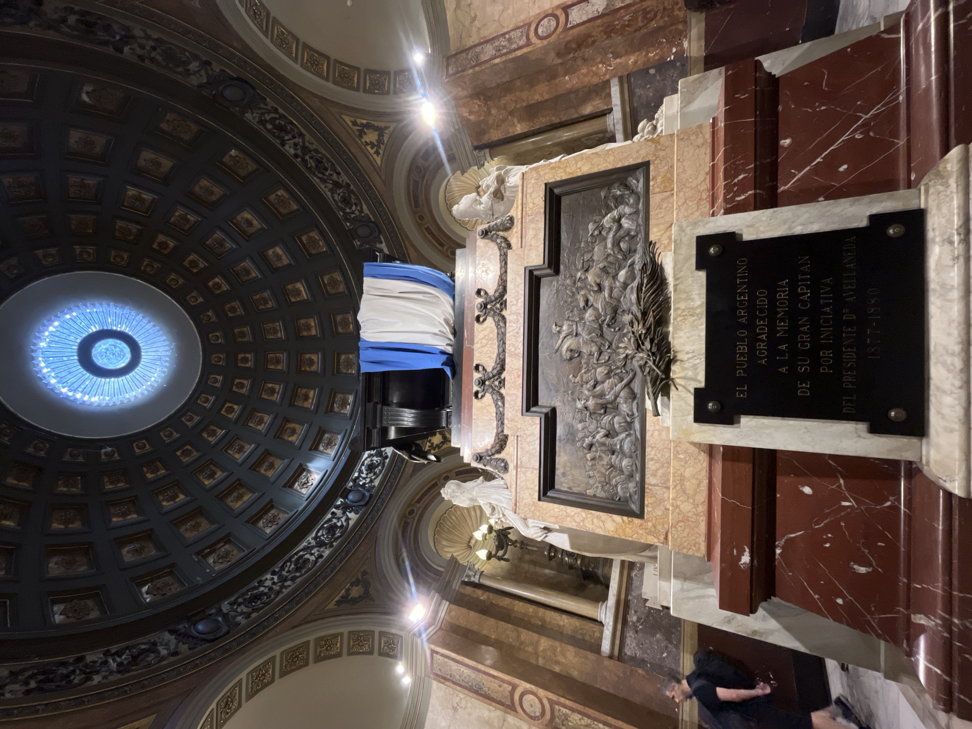 Here's a detailed description of the image:
**General Overview:**
The image depicts a mausoleum located within a large, ornate building. The perspective is from below, looking slightly upwards and towards the side of the mausoleum. A person stands in front of the mausoleum.
**The Mausoleum:**
The mausoleum is constructed from various types of marble, including reddish-brown and tan marble. There is a bronze bas-relief panel, showing a group of riders on horses. A black plaque with white lettering is mounted on the mausoleum. The inscription is in Spanish and reads: "EL PUEBLO ARGENTINO AGRADECIDO A LA MEMORIA DE SU GRAN CAPITAN POR INICIATIVA DEL PRESIDENTE DE AVELLANEDA 1877-1880." A draped cloth is resting on the monument.
**The Building Interior:**
Above the mausoleum is a large, domed ceiling with an elaborate, patterned design. The dome has a central chandelier. The walls and surrounding areas are also decorated with intricate designs and ornamentation.
**Person:**
One person is visible. The individual is standing in front of the mausoleum.
**Environment:**
The location is likely a mausoleum in Argentina, given the inscription. The architecture suggests a significant historical site.