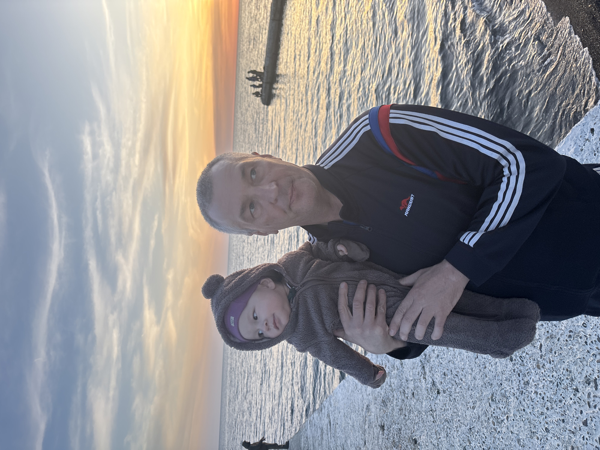 A man holding a baby stands on a concrete structure by the sea. The man is wearing a dark-colored jacket with white stripes and the baby is wearing a brown hooded outfit. The sea is in the background with a breakwater visible in the distance. The sky shows colors of sunset. The location is Sochi, Russia.