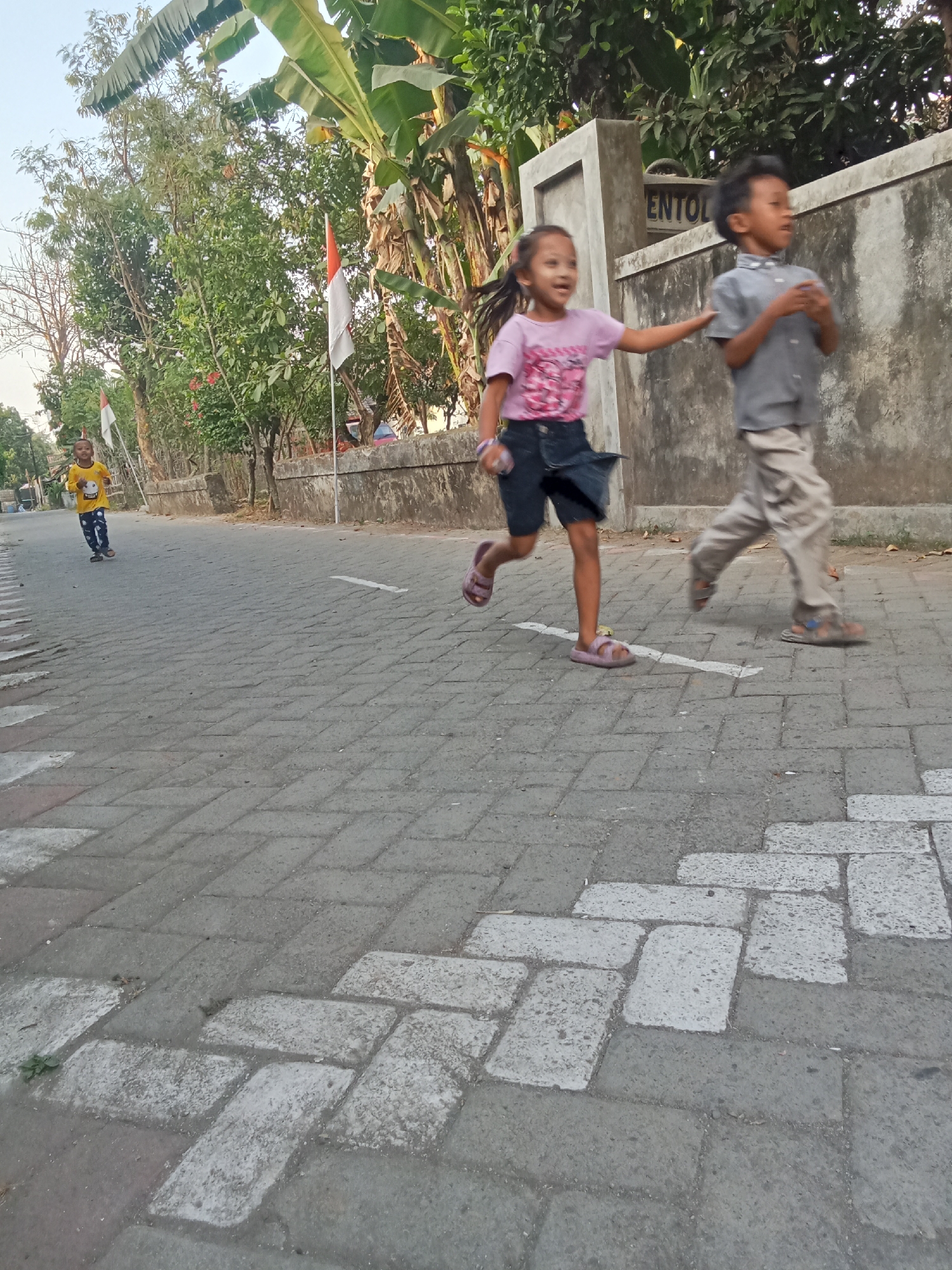 In the city of Keplaksari, Indonesia, a group of children are running on a paved walkway adjacent to a wall. In the foreground, a girl in a pink t-shirt and denim shorts is in motion, her right hand reaching towards a boy in a grey shirt and light-colored pants. The boy is also running, looking to his right. Further down the walkway, a young boy in a yellow t-shirt and patterned pants is running towards the foreground. The environment features lush green trees and foliage on the left side, with a glimpse of a white and orange flag on a pole. The walkway is constructed of interlocking gray pavers with some white markings. A rough, concrete wall runs along the right side of the walkway, with the partial inscription "ENTOL" visible on it. The sky above is a pale blue, suggesting daylight.