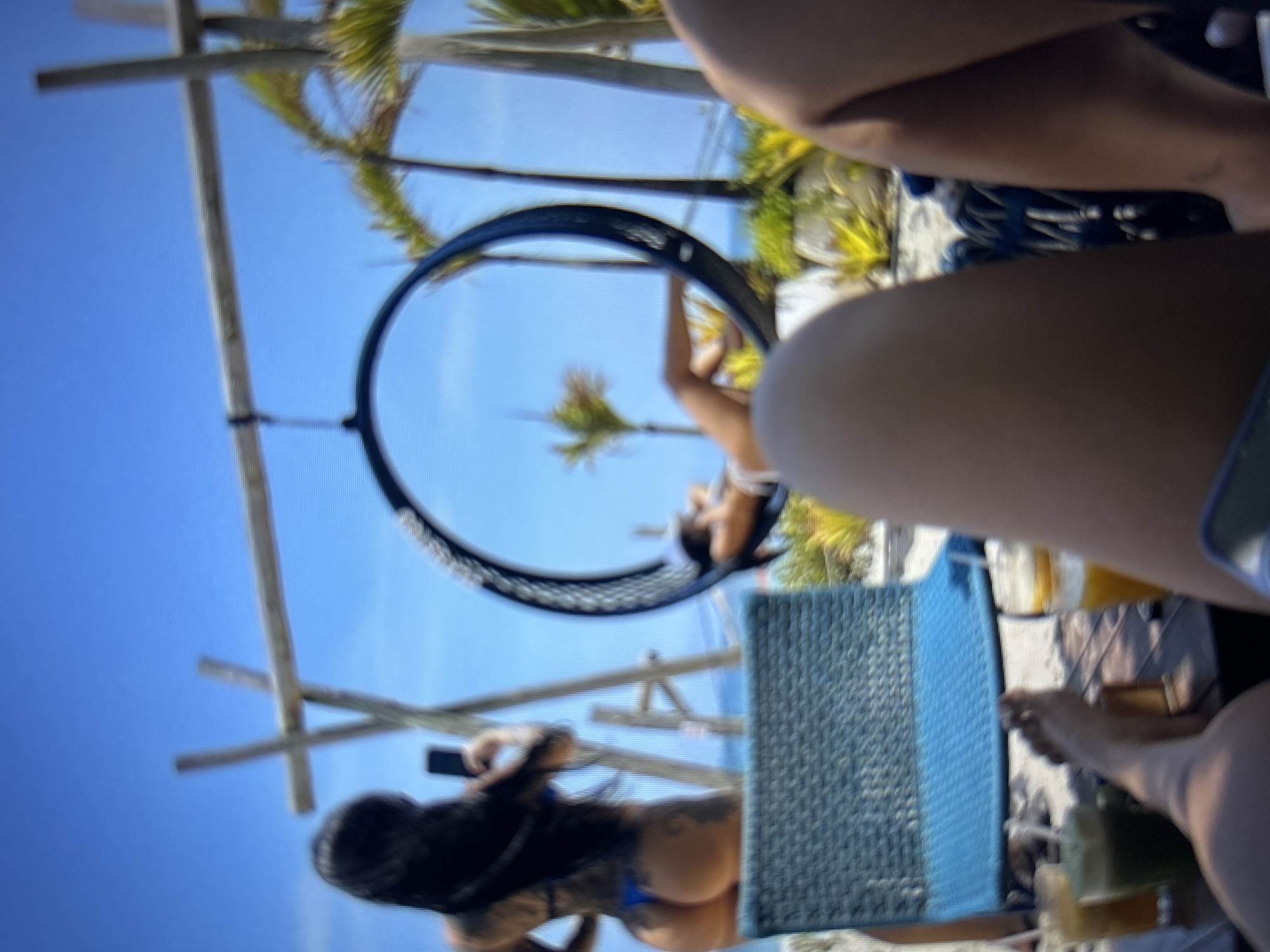 This image depicts an outdoor scene at a beach or resort location in Burgas, Bulgaria, captured from a first-person perspective. The foreground shows the lower legs and bare feet of an individual, along with a wooden table holding at least two beverages in glasses: one green smoothie and one orange-colored drink. A blue woven chair is positioned in the immediate midground.
Further back, a woman with long dark hair and visible tattoos on her back and buttocks is standing, wearing a blue thong bikini bottom. She holds a dark-colored smartphone, appearing to take a photograph or video of another individual. This second individual is partially obscured and is reclining in a large, dark-colored circular swing that is suspended from a wooden structure. The swing has a woven or textured surface.
In the background, a clear blue sky dominates the upper portion of the frame, with the blue ocean visible horizontally. Scattered palm trees and other tropical foliage are present, along with additional wooden support structures. The overall environment suggests a sunny, leisurely setting.
