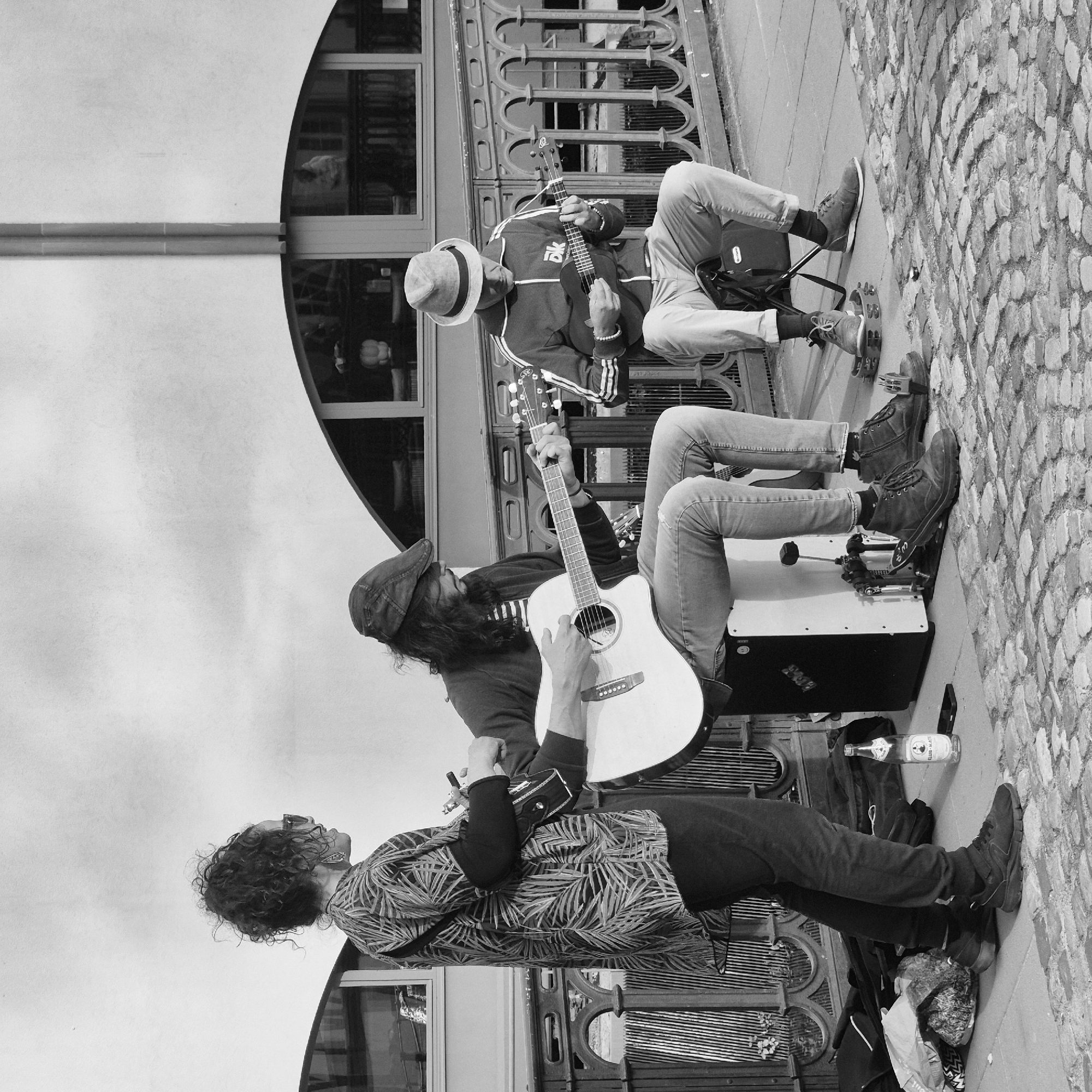 Three individuals are depicted performing music on a cobblestone sidewalk adjacent to an ornate metal bridge railing in Strasbourg, France. The scene is monochromatic.
On the left, a person with curly hair, wearing sunglasses, a patterned short-sleeved shirt over dark long sleeves, and dark trousers, stands holding a small, dark stringed instrument. To their immediate right, a bottle of "CLUB-MATE" and a smartphone rest on the pavement. The central figure, seated on a black and white cajón drum, wears a flat cap, a dark jacket over a striped shirt, and light-colored jeans; they are actively playing a light-colored acoustic guitar. To the right, the third individual, wearing a fedora, a dark track jacket with white stripes, and light-colored trousers, sits on a dark folding stool, playing a dark-colored ukulele. A tambourine is positioned beneath this person's left foot.
In the background, a light-colored building with large, arched-top windows reflects interior details and faint figures. The bridge railing, dark and featuring decorative arch patterns, separates the performers from the background. The ground transitions from a cobblestone surface on the right to a smoother paved area on the left, where various bags and personal items are visible.