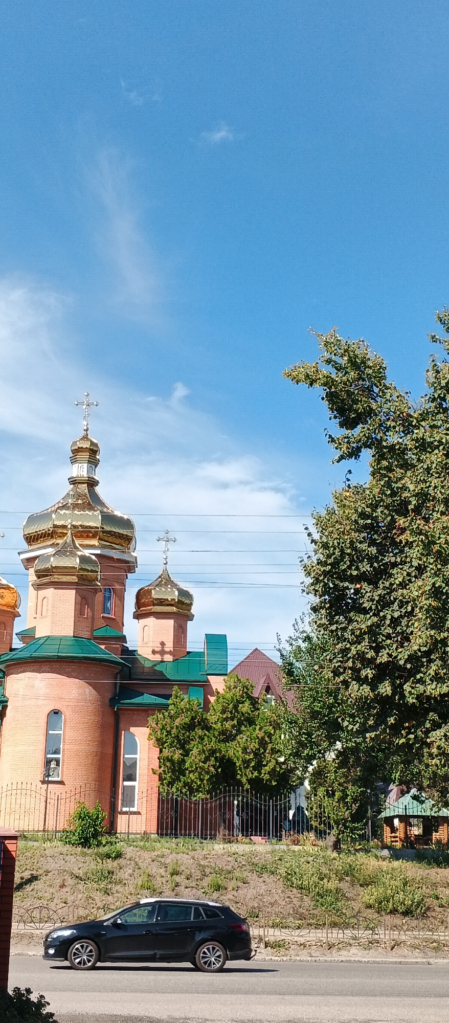 In the city of Smila, Ukraine, a black station wagon is parked on an asphalt road, with its left side facing the camera. The car is positioned in front of a brick building with golden domes and green roofs, resembling a church. The building is surrounded by a black metal fence and a grassy slope with scattered bushes. A large, leafy tree with green foliage is situated to the right of the building, its branches extending towards the blue sky with scattered white clouds. Power lines are visible above the building and tree. The overall scene depicts a sunny day in a Ukrainian city.
