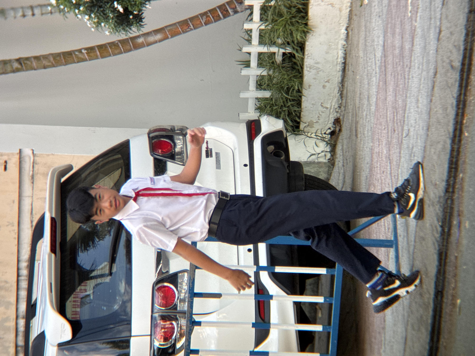 A young male wearing a white short-sleeved collared shirt with a red tie and dark trousers walks past a white SUV in Nha Trang, Vietnam. The SUV has its rear end visible, including taillights and a spoiler. The background shows a light-colored wall and a palm tree. A small white picket fence and greenery are also present. The ground is paved with concrete and some sand. The image appears to be taken outdoors during the day.