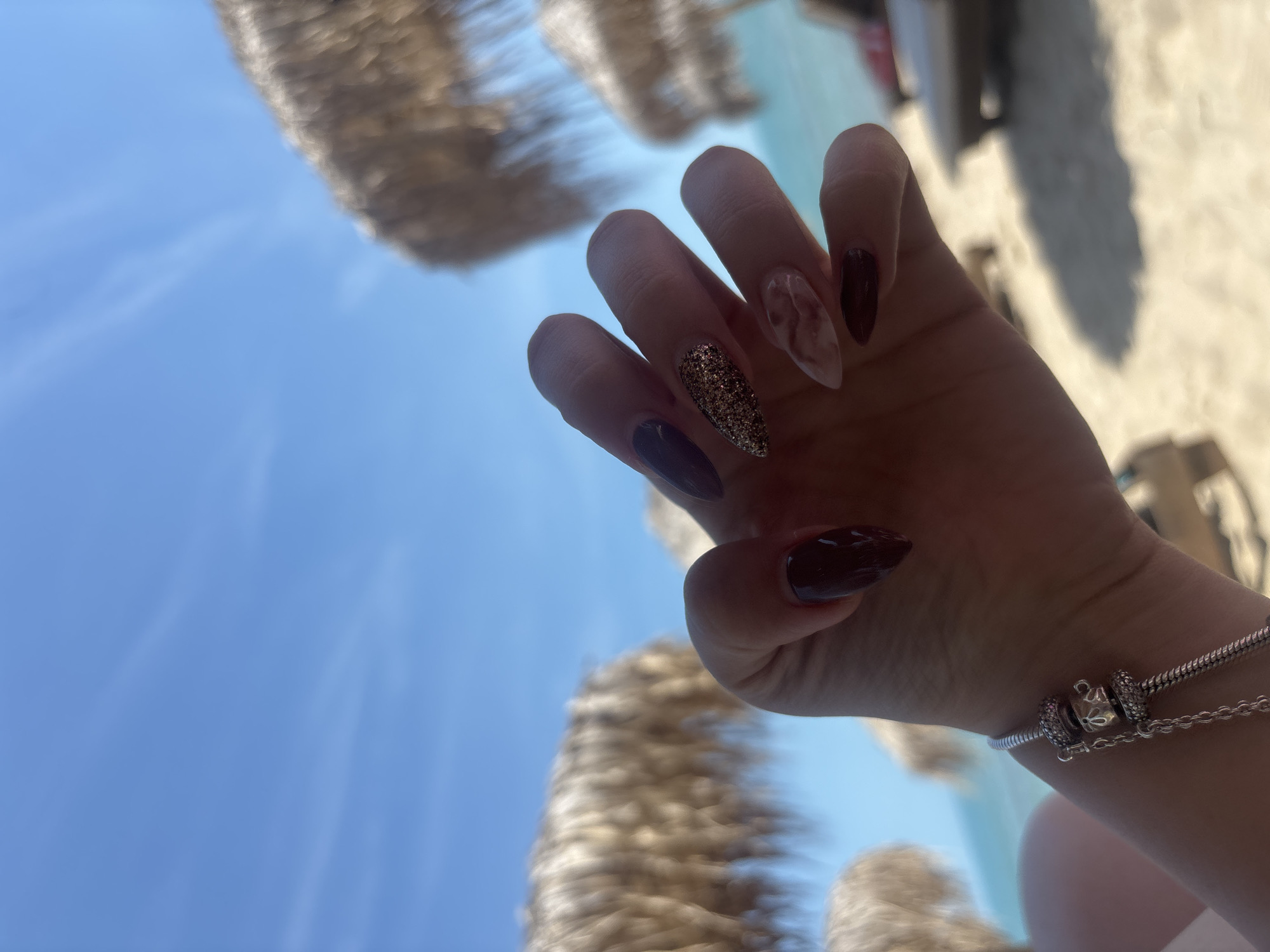 A close-up view depicts a person's left hand, angled upwards, against a blurred backdrop of a sunny beach in Kranevo, Bulgaria. The hand exhibits long, almond-shaped artificial nails: the thumb, index, and pinky fingers are painted solid dark burgundy; the middle finger displays a gold glitter finish; and the ring finger features a light beige base with a reddish-brown marbled design. A silver-colored charm bracelet, consistent with a Pandora style, is visible on the wrist, featuring a snake chain and at least two detailed charms, one pavé-set and another with an openwork design. The background reveals a clear, bright blue sky with faint cirrus clouds, a turquoise sea line, light-colored sand, and several out-of-focus thatched beach umbrellas and lounge chairs, indicative of a coastal resort setting. The lighting suggests bright daylight conditions.