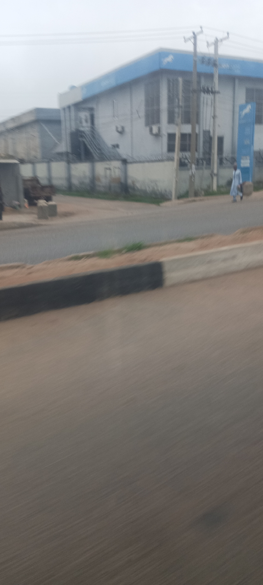 The image captures a street scene in Jos, Nigeria, taken from a moving vehicle, evidenced by the motion blur in the foreground. A two-lane asphalt road with a concrete median occupies the lower and middle sections of the frame.
On the right side of the road, a person wearing light-colored, long clothing (possibly a traditional garment) is walking, carrying a dark object in their left hand. Behind them, a prominent two-story building with a light grey facade features a distinctive blue rooftop trim and a vertical blue sign. Both the trim and the sign display a white horse logo and the text "STANBIC IBTC," indicating a branch of Stanbic IBTC Bank. Air conditioning units are visible on the building's exterior, and tall, vertical window structures line its face.
In front of the bank building stands a tall utility pole with multiple crossarms and electrical wires. A white concrete fence topped with barbed wire runs along the roadside, separating the bank property from the street. Another, less defined building is partially visible behind the Stanbic IBTC structure to the left. The sky is uniformly overcast or hazy, casting a diffused light over the entire scene, suggesting a cloudy day or early morning/late afternoon conditions.