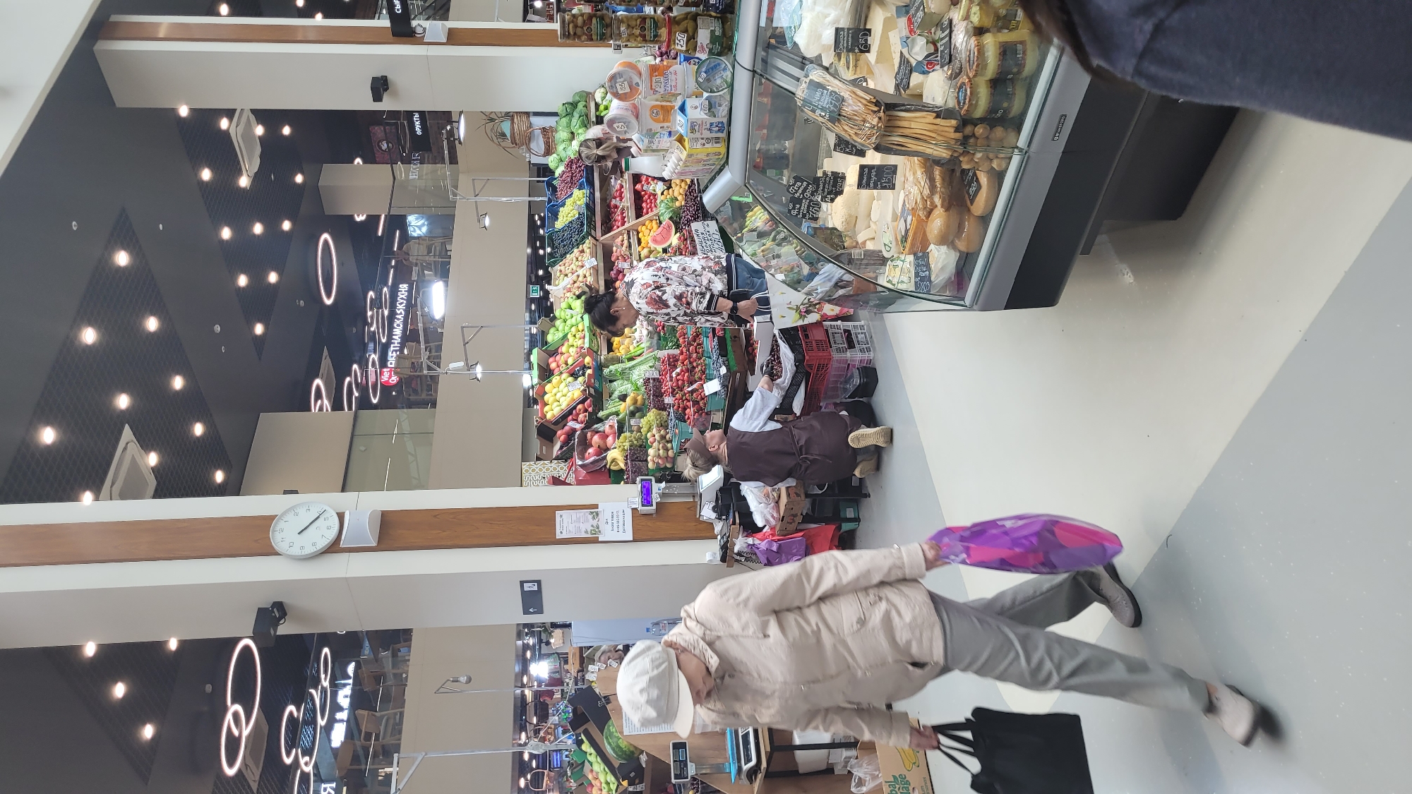 In a supermarket, a woman wearing a light-colored hat and jacket walks past a fruit and vegetable display. She carries a black tote bag in her left hand and a purple and pink shopping bag in her right. To her right, a person in a brown apron kneels to arrange fruit in crates, while another person stands in front of the display. Beyond them, a refrigerated case displays various cheeses and food items. The scene is illuminated by overhead lights and features a large clock on a pillar.