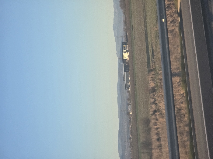 The scene depicts a rural landscape near Diadovo, Bulgaria, observed from a road. In the immediate foreground, a dark grey metal guardrail spans horizontally, positioned above a segment of a paved road with visible white lane markings. Beyond the guardrail, the midground consists of extensive fields displaying brown and green vegetation, interspersed with patches of dry, reedy growth and sparse, bare trees or bushes. In the distant background, a prominent industrial-style building complex is visible within these fields, composed of a large, dark grey structure with an attached lighter-colored section. A range of low, undulating mountains forms the furthest horizon. The upper two-thirds of the image are occupied by a clear, gradient pale blue sky, indicative of daylight. No individuals or discernible activities are present.