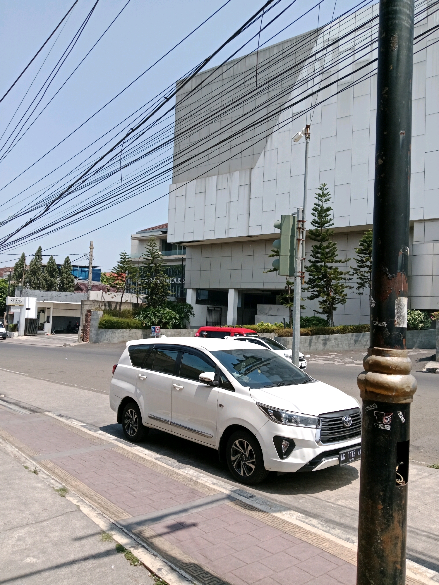In the city of Cianjur, Indonesia, on a sunny day, a white Toyota Innova is parked along a sidewalk. The vehicle, with license plate AG 1172 MX, faces towards the right of the frame. The sidewalk is paved with reddish-brown bricks and bordered by concrete. Beyond the sidewalk, a road runs parallel to the parked car, with another vehicle, a red car, partially visible in the background. The scene is framed by a tall, dark lamppost on the right, covered in stickers and showing signs of wear. Overhead, a dense network of electrical wires crisscrosses the sky, leading towards a modern, multi-story building with a white, textured facade. The building's architecture is contemporary, with large, rectangular panels. Several evergreen trees are planted in front of the building, adding a touch of greenery to the urban landscape. A traffic light is visible on a pole near the trees. The sky is clear and blue, indicating fair weather.