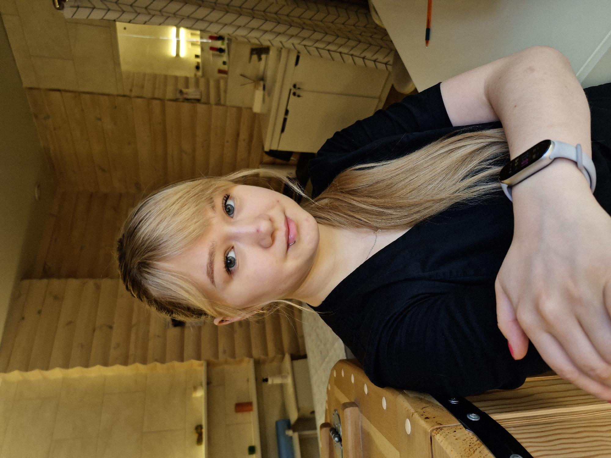 A young woman with blonde hair and blue eyes is looking at the camera. She has a lip piercing on the left side. She is wearing a black shirt and a smartwatch on her left wrist. She is indoors. The background includes wood paneling, a white cabinet, and a tiled area. It is located in the city of Chaikovsky, Russia.