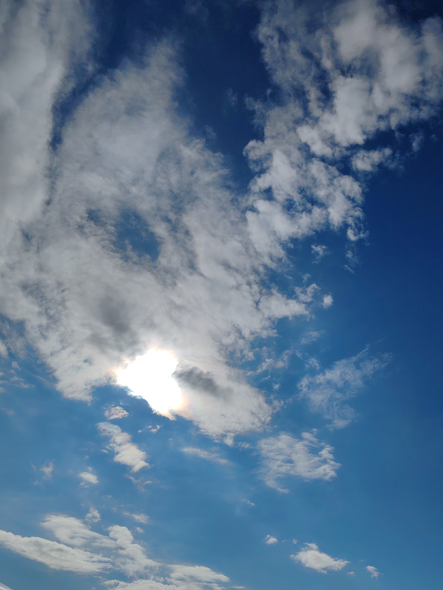 The image shows a bright, sunny day with a clear blue sky and scattered white clouds. The sun is partially obscured by a large, wispy cloud formation in the upper left quadrant, casting a bright glow and some subtle rainbow hues. Smaller, fluffy clouds are distributed across the sky, with some appearing lower in the frame. The scene is a generic sky view and does not contain any specific features that would indicate its location within the city of Чухлэм, Russia, or any other city. There are no people, subjects, objects, or actions depicted in the image.