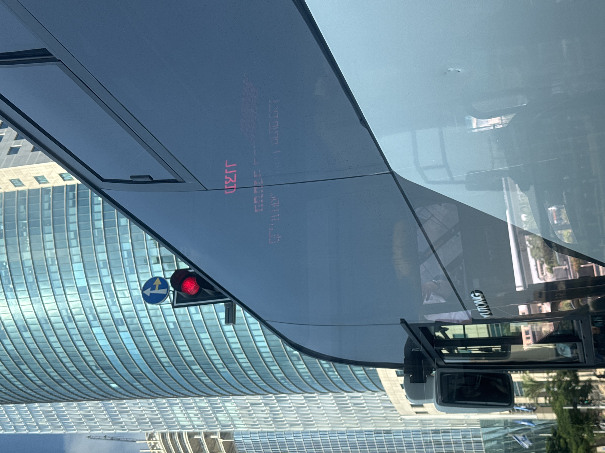 The image captures a street scene in Tel Aviv, Israel, with a focus on a bus and modern skyscrapers. The bus, a Yutong model, has its digital display illuminated with text in red Hebrew characters. The larger text reads "יציאה" (Yetziah), meaning "Exit". Below this, a second line of text appears to be a route number or destination, possibly "702 - 460". Reflections on the bus's dark, tinted windows show buildings and the sky.
To the left of the bus, a traffic signal displays a red light. Above it, a blue circular sign with a white arrow indicates a mandatory right turn. Dominating the background are tall, modern buildings with reflective glass facades, characteristic of Tel Aviv's business districts. One particularly prominent building is a curved skyscraper with a blue-tinted glass exterior. Another, taller building to the far left is under construction, with its skeletal framework visible. The sky is partly cloudy. The overall impression is a dynamic urban environment.
