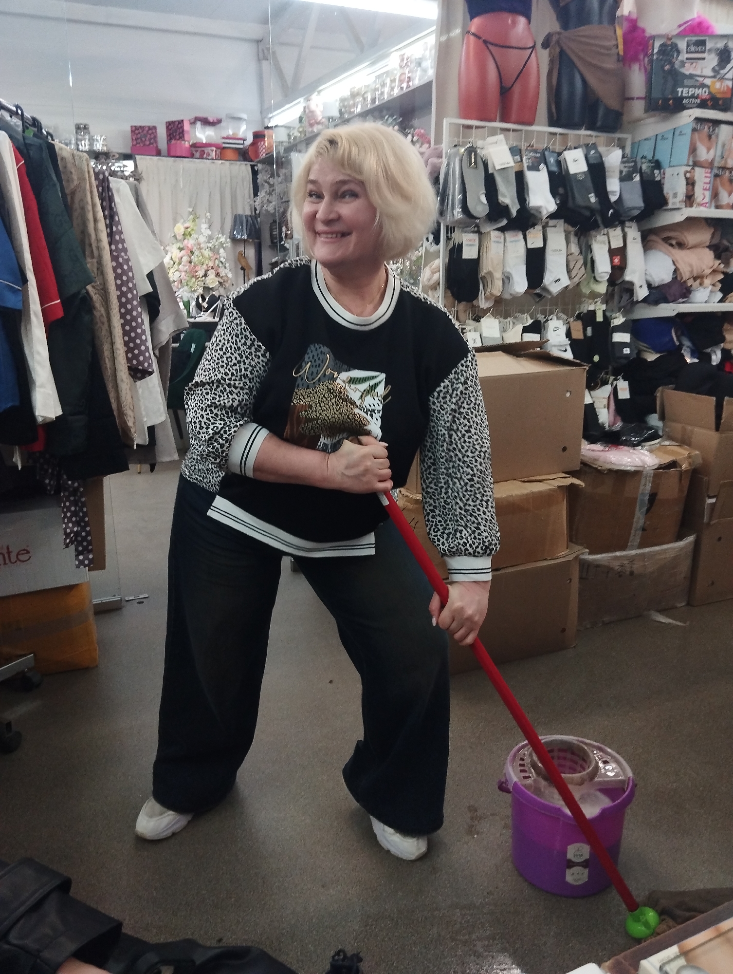 The image depicts a woman holding a mop inside a retail store. The woman has short blonde hair and is wearing a black and leopard print sweatshirt and black pants. She's holding a red-handled mop next to a purple bucket with a wringer. Behind her are racks of clothing, shelves filled with merchandise (including socks and undergarments), and stacks of cardboard boxes. The store appears to be located in Votkinsk, Russia, based on product packaging visible in the background.