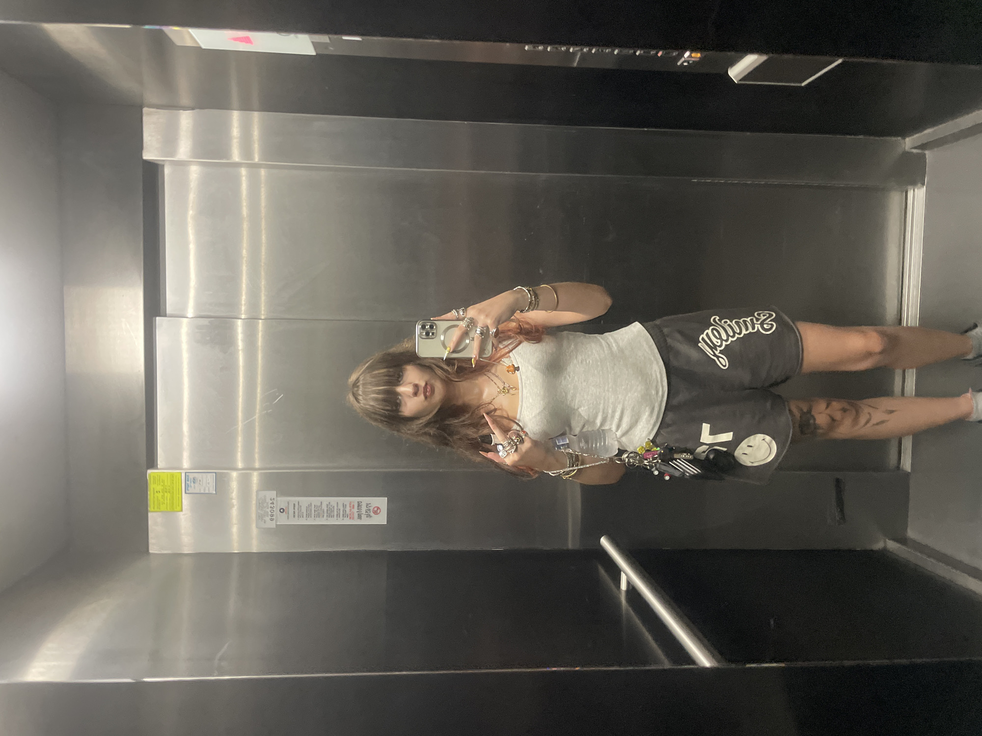 A young woman with brown hair and bangs is taking a selfie in an elevator. She is wearing a grey tank top, dark shorts with a smiley face logo and script text, and several rings and bracelets. She is holding a water bottle and a keychain in her left hand. The elevator has stainless steel walls and a black panel on the left side with a handrail. There is a sticker on the wall behind her with Hebrew text and a yellow rectangle. The scene appears to be in Raanana, Israel, based on the context provided.