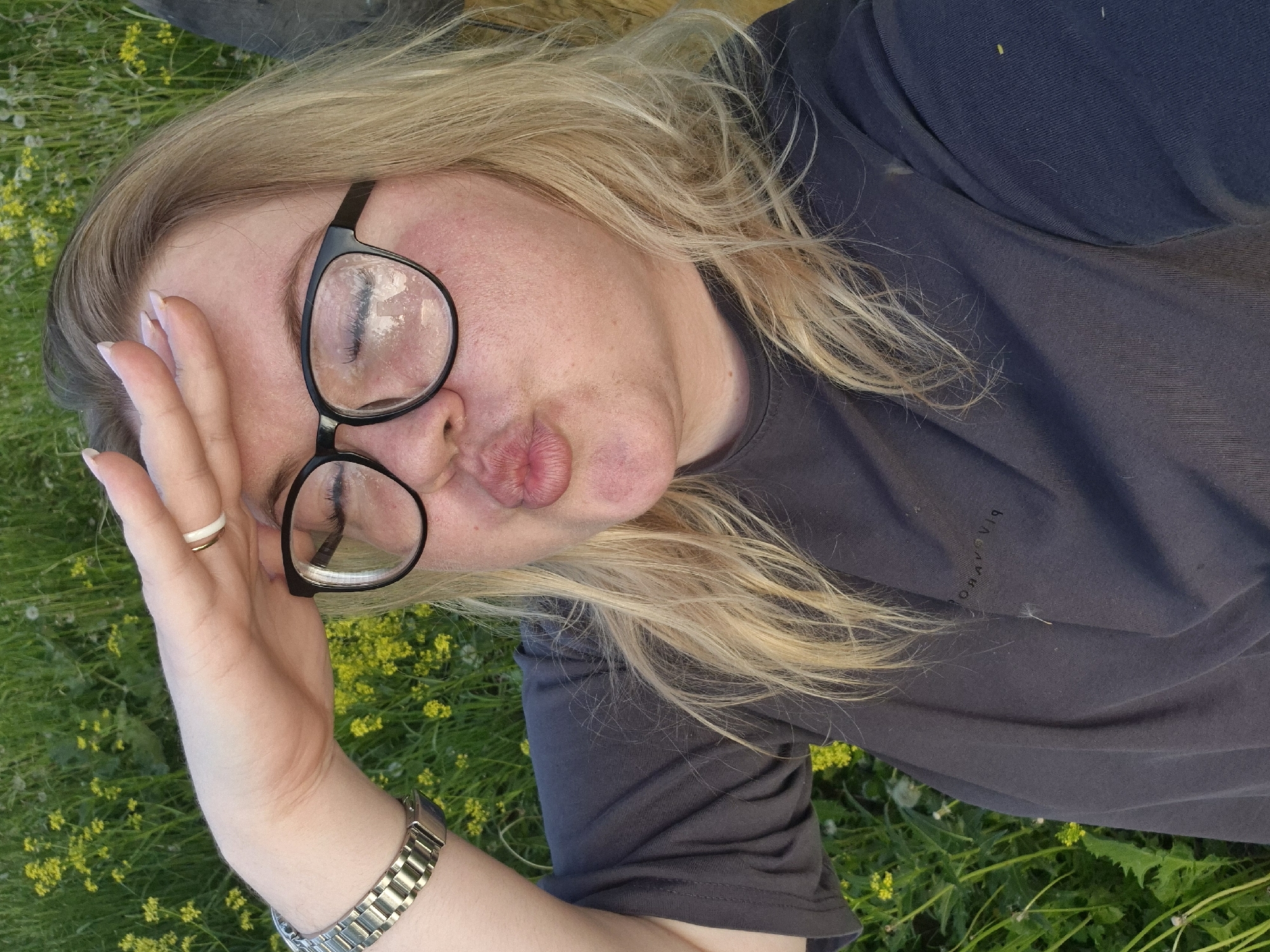 The image, taken in Фоки, Russia, features a medium close-up of a person, likely a woman, lying down outdoors in a field of green grass with small yellow flowers. She has long, wavy blonde hair and is wearing a dark grey or charcoal t-shirt. Her face is visible, adorned with round, black-framed glasses whose lenses appear smudged or condensed, obscuring her eyes. Her lips are pursed in a "kiss face" or "duck face" expression.
Her left hand is raised to her forehead or temple, with her fingers lightly resting there. She wears a two-tone ring (white and gold/cream) on her left ring finger and a metallic, silver-link watch on her left wrist. The background consists entirely of lush green grass speckled with numerous bright yellow wildflowers, suggesting a natural outdoor setting like a meadow or garden, under natural daylight.
On the front of her dark t-shirt, near her right shoulder from the viewer's perspective, there is stylized text that appears inverted and possibly mirrored. It resembles characters such as "ЯІЯΔЯОЯq", which, when corrected, could spell out a word like "BLACKDROP". The overall impression is one of a casual, relaxed, and possibly playful moment outdoors.