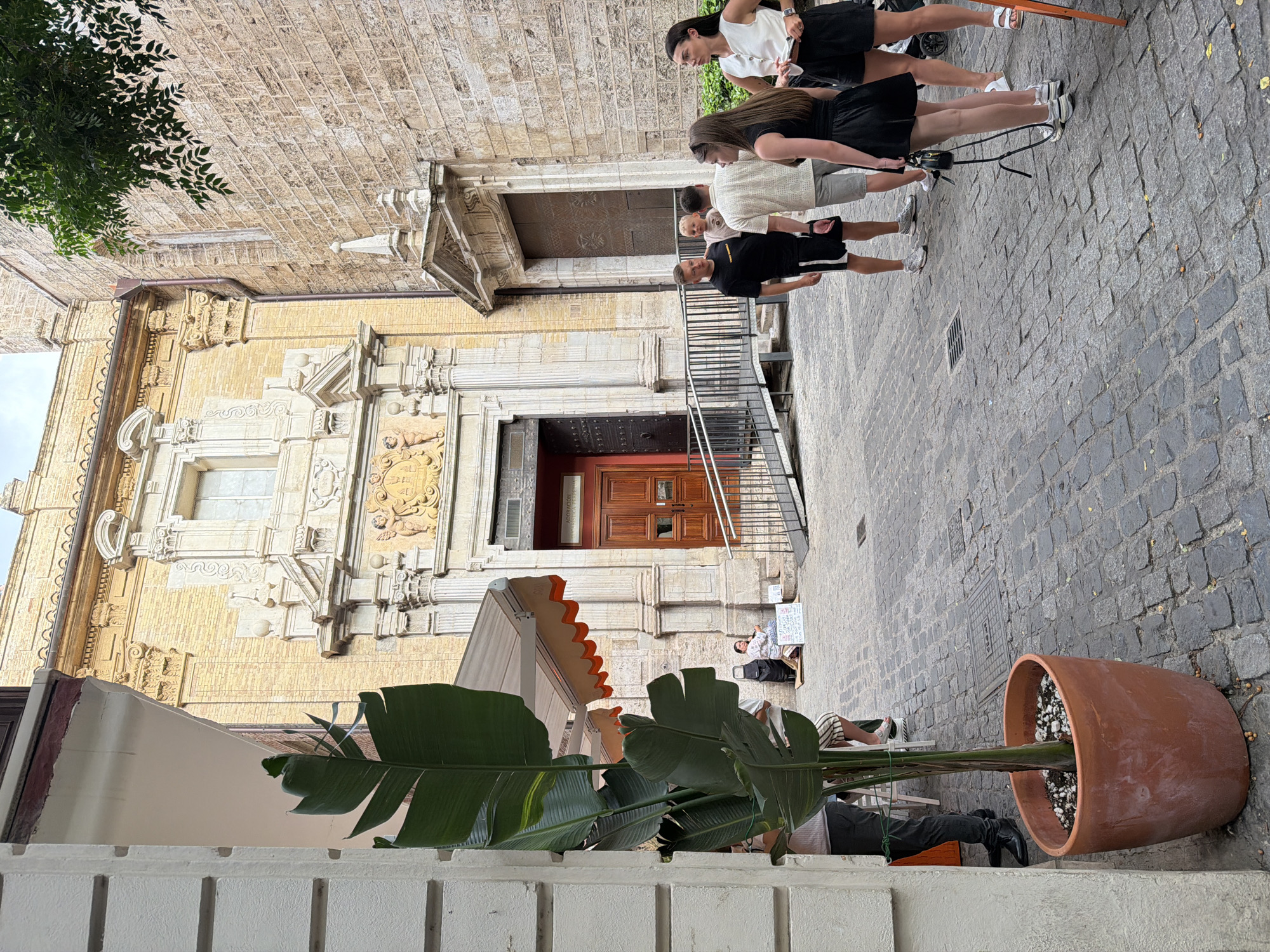 In an urban courtyard paved with cobblestones, a group of people are gathered near the entrance to a building with ornate architecture. A large potted plant with broad green leaves stands in the foreground on the left. The building's facade is constructed of yellow brick and features intricate stone carvings and a large wooden double door, possibly an entrance to a church or historical site. To the right of the main entrance, another stone-clad building with a similar architectural style is visible.
Several individuals are interacting in the middle ground. A man in a black t-shirt and shorts is looking towards the left, while a baby stands between him and another man wearing a light-colored shirt and shorts. Two women are also present; one in a black dress is holding a small handbag and looking towards the right, and another woman in a white sleeveless top and black shorts is standing nearby.
Further back, near the building's entrance, a woman sits on the ground with a cart and some signs. The environment suggests a public outdoor space in a European city, likely a historic district. The lighting indicates it is daytime.