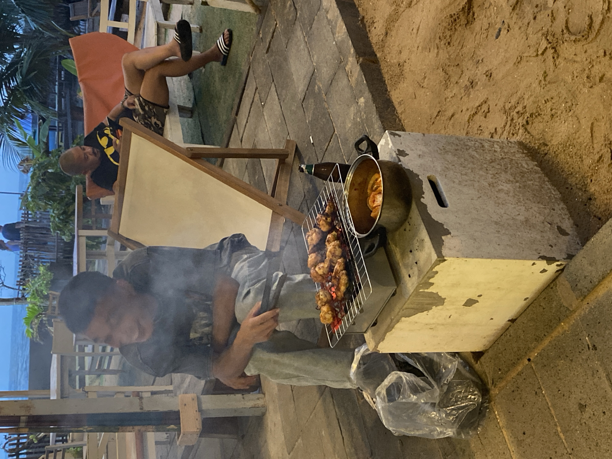 A young man sits on a concrete block at an outdoor seating area, tending to a small charcoal grill laden with grilled chicken skewers. He is wearing a dark t-shirt and light-colored pants, and he appears to be smiling. Smoke rises from the grill, obscuring his face slightly. To his right, a metal pot containing what appears to be more cooked food sits on the concrete block, next to a glass bottle. In the background, another man, wearing a black t-shirt with a Batman logo and camouflage shorts, sits on a bench and looks at his phone. He is wearing flip-flops. The seating area is paved with stone tiles and sand, and tables and chairs are visible in the background, suggesting a restaurant or beachside establishment. Palm trees and a wooden fence are also visible, indicating a tropical location. The overall scene suggests a casual dining experience. The location is Mũi Né, Vietnam, known for its beaches and seafood restaurants.