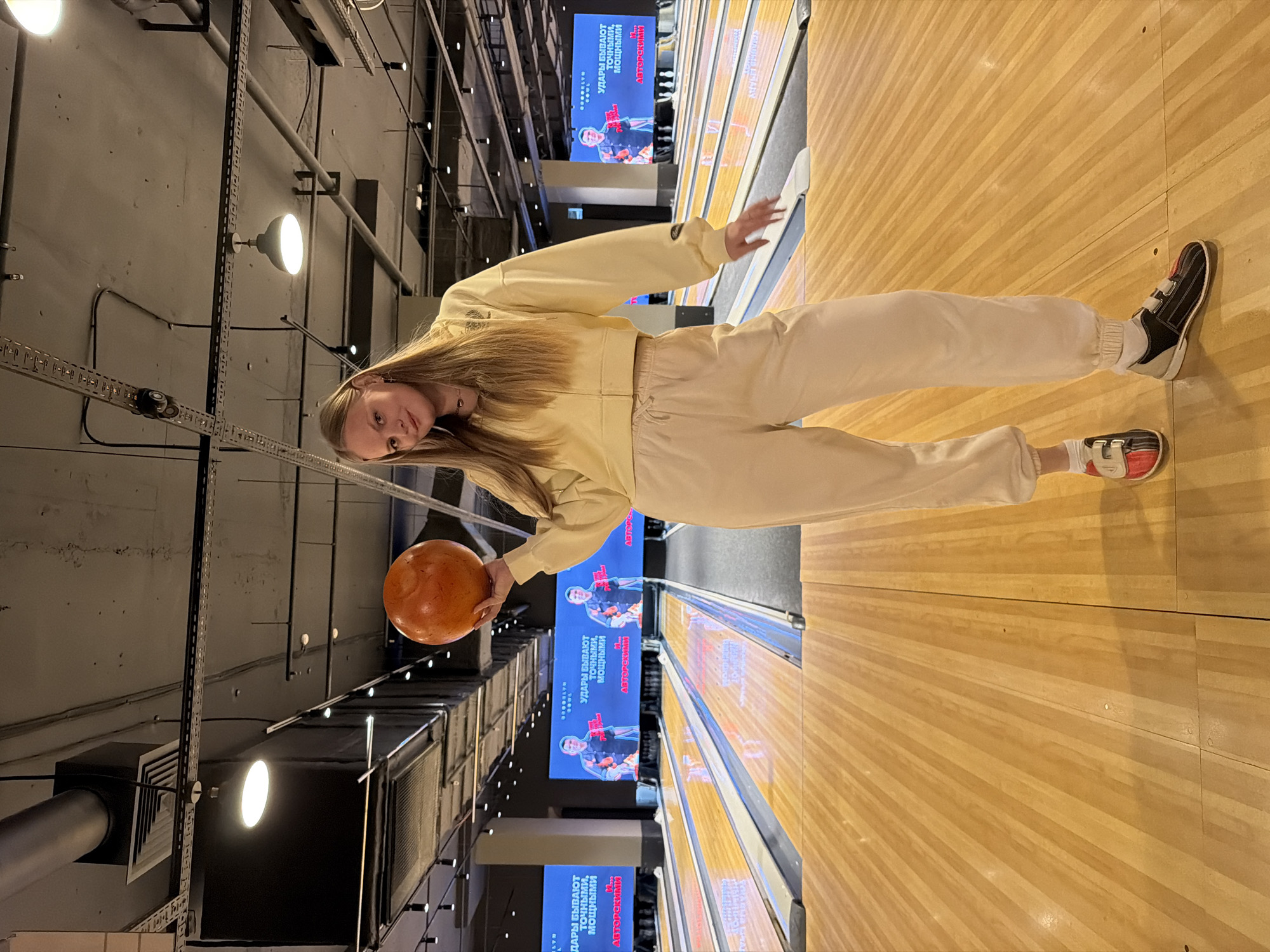A young woman is captured in a bowling alley in Izhevsk, Russia, holding an orange bowling ball, appearing to pose or prepare for her turn. She has long, straight blonde hair and is dressed in a casual light yellow zip-up hoodie and matching light-colored sweatpants. Her feet are clad in two-toned bowling shoes, one predominantly black and white, the other black, white, and red.
The setting is an indoor bowling facility, characterized by highly polished wooden bowling lanes stretching into the background. The ceiling above features an industrial aesthetic with exposed pipes, ventilation ducts, and a grid system supporting numerous artificial lights, some emitting a bright glow.
In the background, several large screens display what appears to be a score or advertisement. Visible text in Cyrillic reads: "УДАРЫ БЫВАЮТ точными, мощными" (Hits are accurate, powerful) and "АВТОРСКИМИ" (By authors/author's). One screen also shows a "BROOKLYN BOWL" logo, indicating the venue name. The overall lighting suggests an evening or nighttime activity. The woman's expression is neutral as she looks towards the camera.