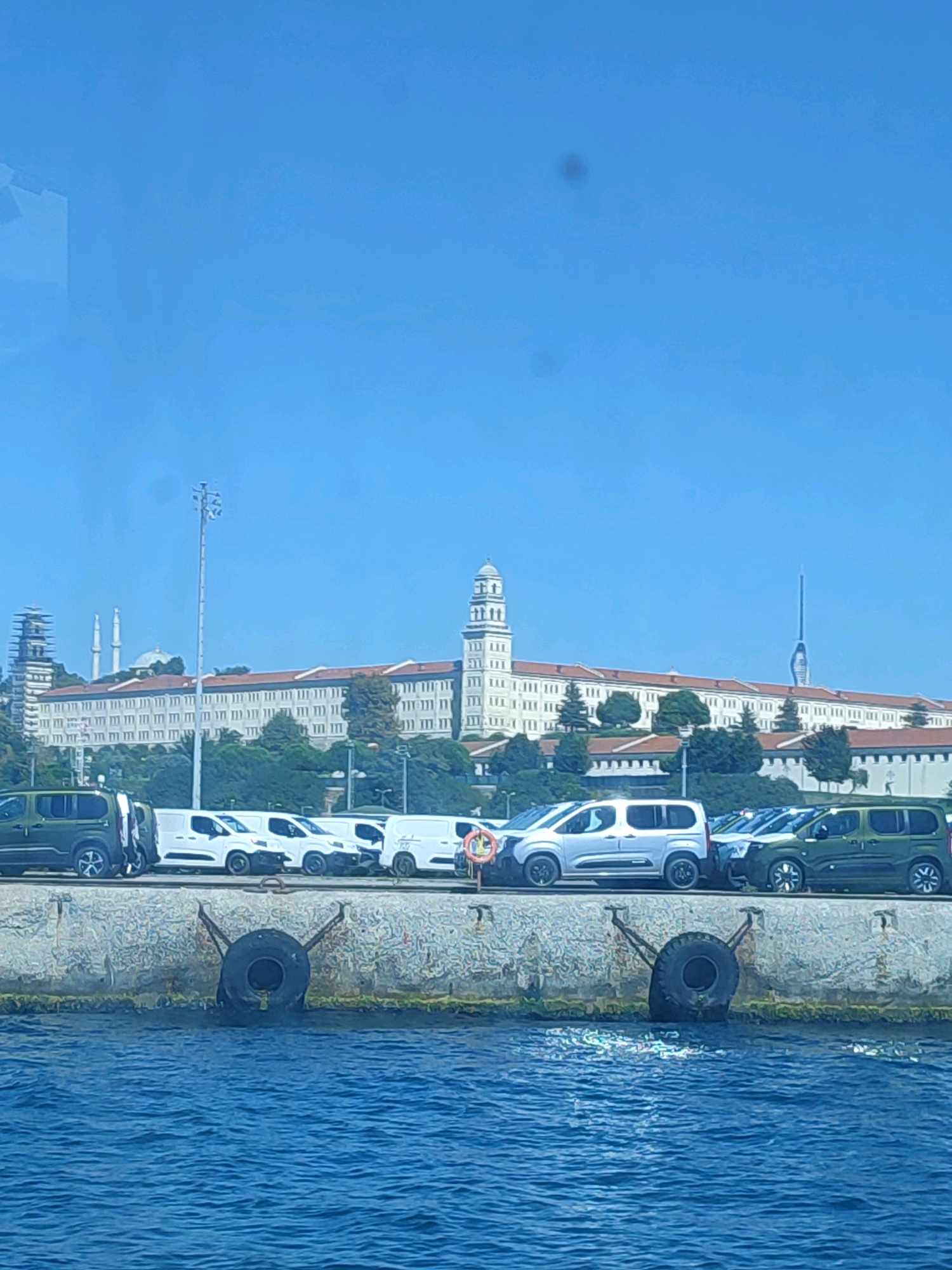 The image captures a waterfront scene in Üsküdar, Turkey, dominated by a large, ornate building complex with a central tower. The building appears to be a historical institution or university, characterized by its long, white facade, red-tiled roof, and numerous windows. Two minarets are visible to the left of the main structure, suggesting a connection to Islamic architecture. A tall, modern communication tower stands to the right of the building. In the foreground, a concrete seawall with two large truck tires used as fenders separates the water from a row of parked vehicles, including several white vans and two green SUVs. The water is a vibrant blue, with small ripples on its surface. The sky is clear and bright blue, indicating a sunny day. The overall impression is a blend of historical architecture and modern infrastructure, set against the backdrop of the Bosphorus Strait.