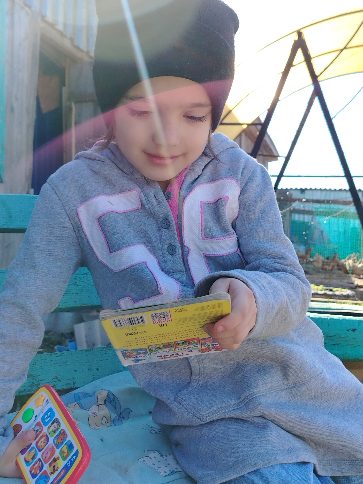 A young girl wearing a black beanie and a gray hooded sweatshirt with a "56" design is sitting on a turquoise bench outdoors in Гожан, Russia. She is holding a small, yellow book in her hands and appears to be reading it. Beside her on the bench is a toy phone with colorful animal icons. The background shows a fence, vegetation, and what appears to be a frame structure with a translucent cover, possibly a greenhouse or outdoor covering. The overall impression is that she is outdoors in a yard or garden area. The image has a lens flare effect near the girl's head.