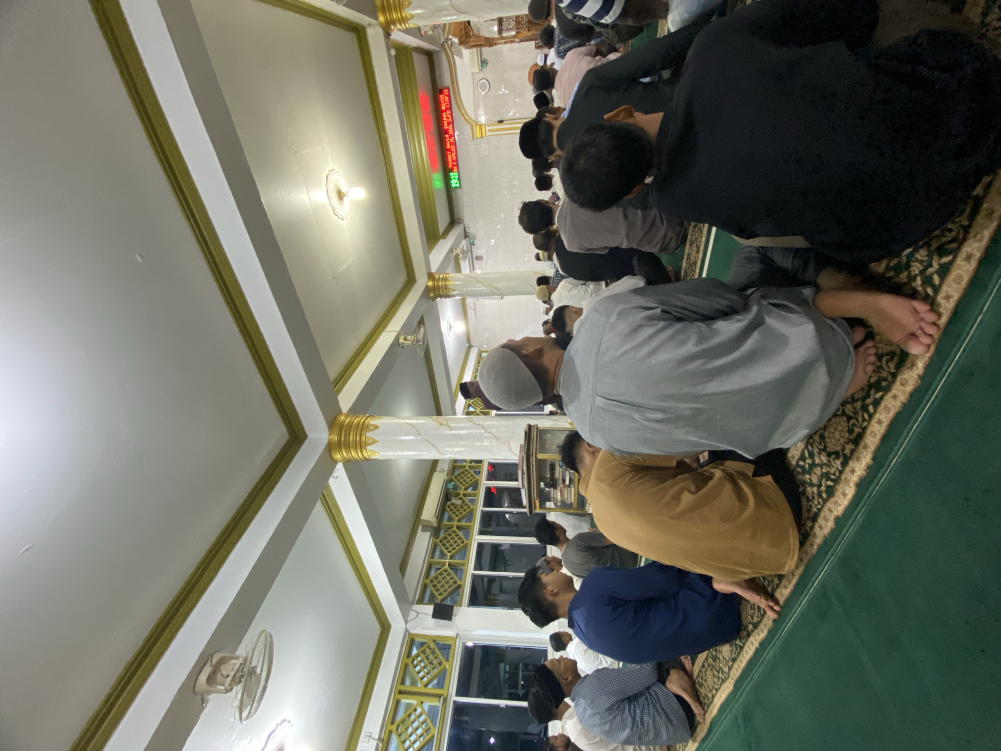 Men are praying in congregation inside a mosque in Lhokseumawe, Indonesia. They are kneeling on a green carpet with an ornate border, aligned in rows, some wearing head coverings. A digital clock displays information in red lettering. The mosque has white walls and pillars with gold accents and windows with patterned bars. A ceiling fan is visible, along with lighting fixtures.