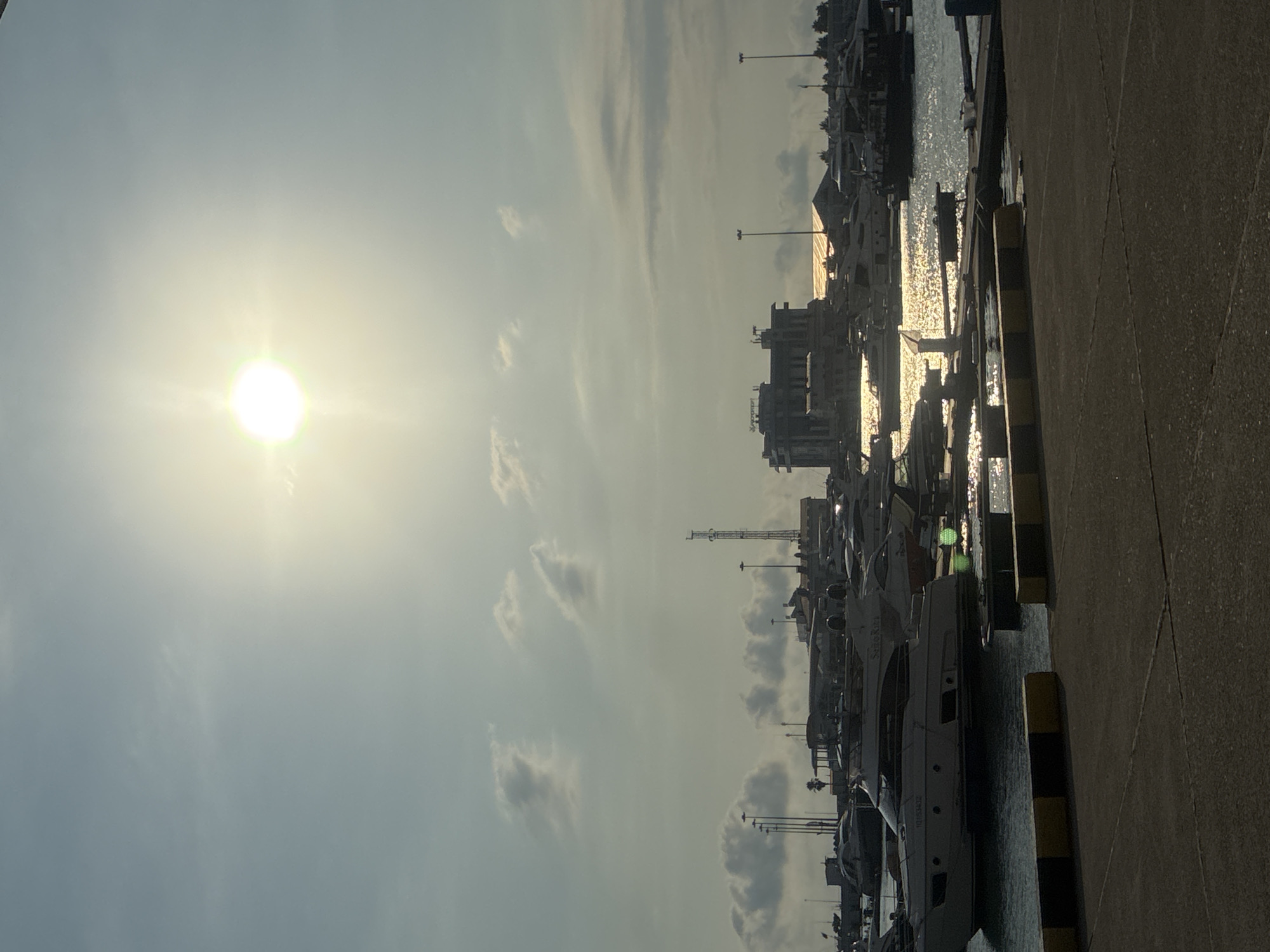 The image depicts a marina in Sochi, Russia, likely the port area. The sun is low in the sky, casting a bright glare on the water and illuminating a cluster of buildings and a radio tower in the background. Several yachts and smaller boats are docked in the harbor, some partially obscured by the glare. The foreground shows a paved walkway with yellow and black striped barriers along the edge of the water. The sky is a pale blue with scattered clouds. There are no people visible in the image.