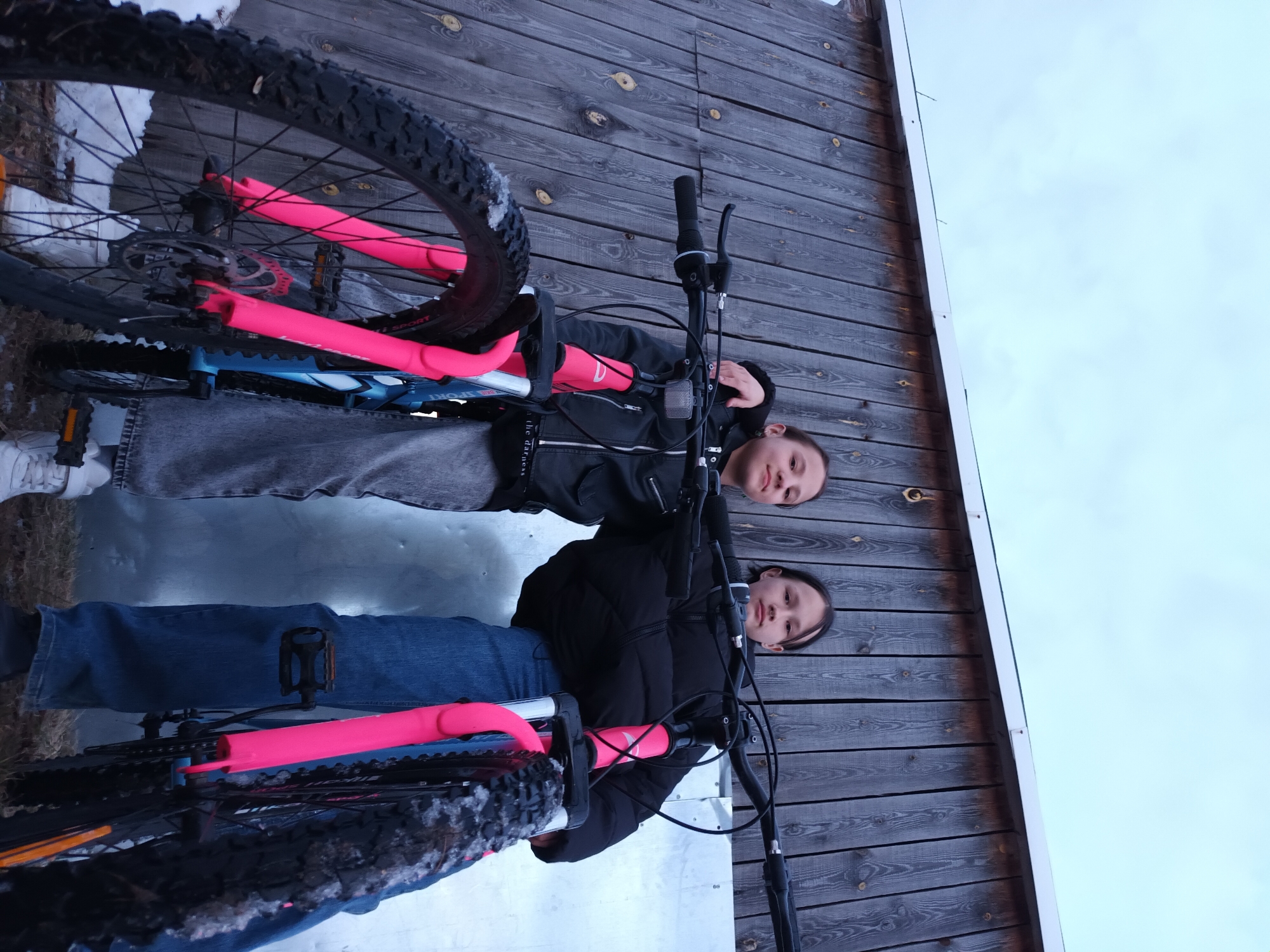 Two individuals, likely adolescent females, pose outdoors in Ольховка, Russia, with two bicycles. The scene is captured from a low-angle perspective, looking upwards.
The individual on the left wears a black leather-style jacket, grey jeans, and white sneakers. Her right arm is draped over the shoulder of the individual on the right. She grips the handlebars of the bicycle positioned closer to the left.
The individual on the right wears a black puffer jacket, blue jeans, and dark shoes. She holds the handlebars of the bicycle positioned to the right.
Both bicycles feature bright pink front forks and blue frames. They are equipped with black knobby tires, some of which exhibit visible snow or ice adhering to the tread and forks, suggesting cold or wintry conditions. Disc brakes are visible on the front wheel of the left bicycle.
The background consists of a weathered, vertically paneled wooden wall on the left, and a light-colored, possibly corrugated metal sheet wall or fence on the right. The ground in the foreground shows patches of snow and dark earth or vegetation. An overcast, light grey sky is visible above the background structures.