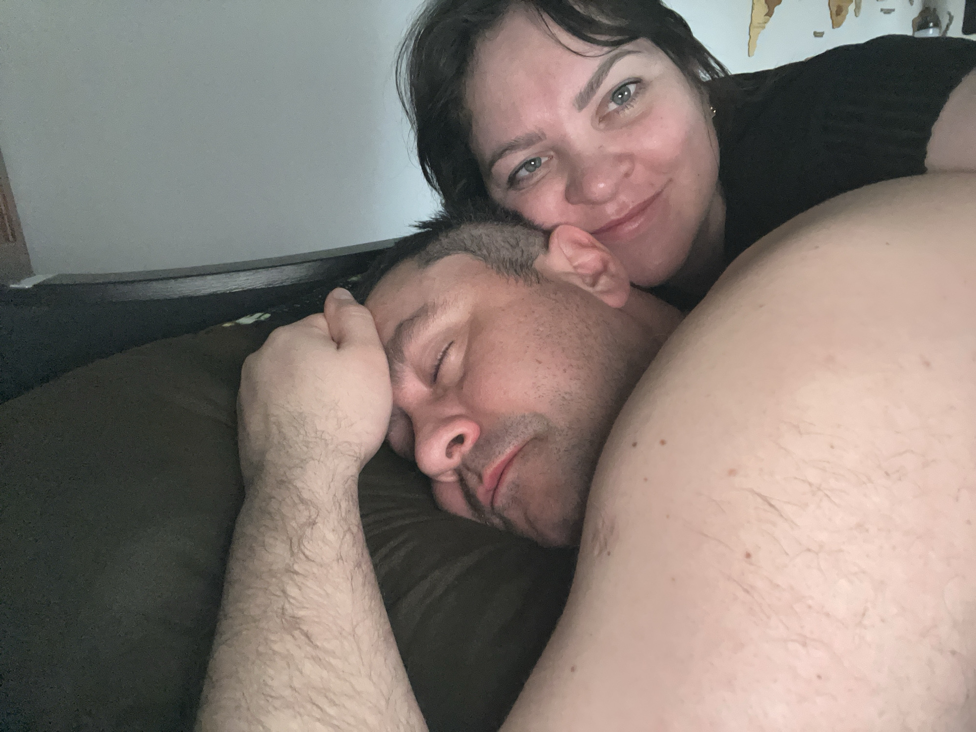 A woman and a man are lying in bed. The woman is awake, smiling, and wearing a black top. The man is sleeping with his eyes closed and his arm raised. The location is unknown and there is no indication of Perm, Russia.