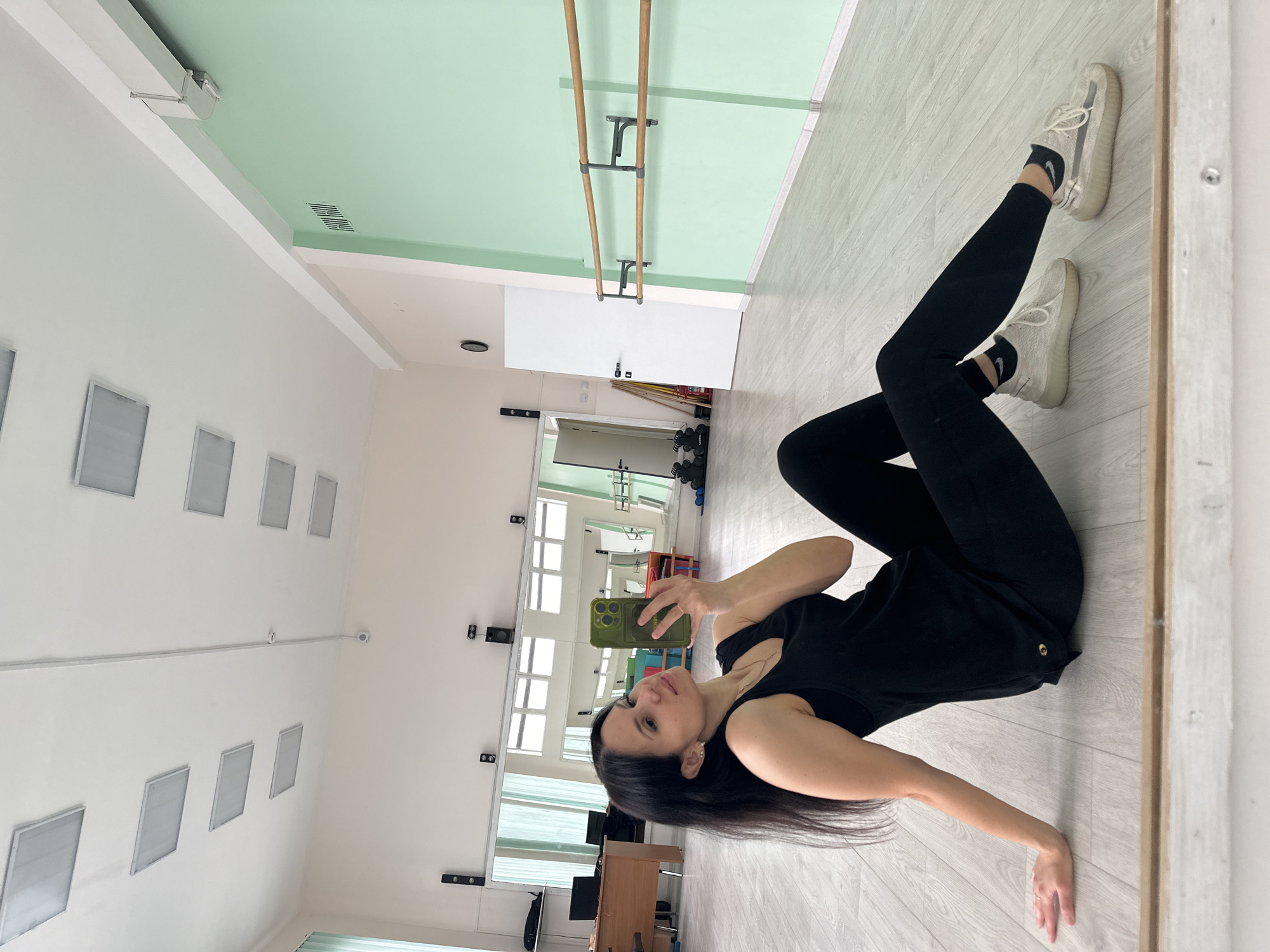 A woman lies on her side on a light grey wood floor in a room with a light green and white ceiling, and white walls. She is wearing a black tank top, black leggings, and off-white sneakers with black socks. She holds a green mobile phone in her right hand to take a selfie in a mirror. The room appears to be a dance studio, with a ballet barre on the wall. There are several evenly spaced, square vents on the left wall. The mirror reflects more of the room, including windows, a desk, and other exercise equipment. Based on the information provided, it is unknown if the location is in the city of Фоки, Russia.