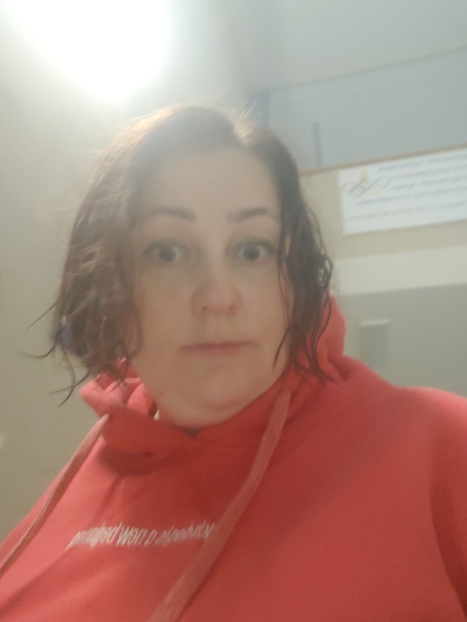 A woman with short, dark curly hair and a red hoodie is looking directly at the camera with a slightly surprised expression. The hoodie has white text embroidered on the left side. The background is blurred, but appears to be an indoor setting with a light-colored wall. A bulletin board is visible in the upper right corner. The lighting is bright, possibly from an overhead light. There is nothing to indicate this image was taken in the city of Markovskii, Russia.