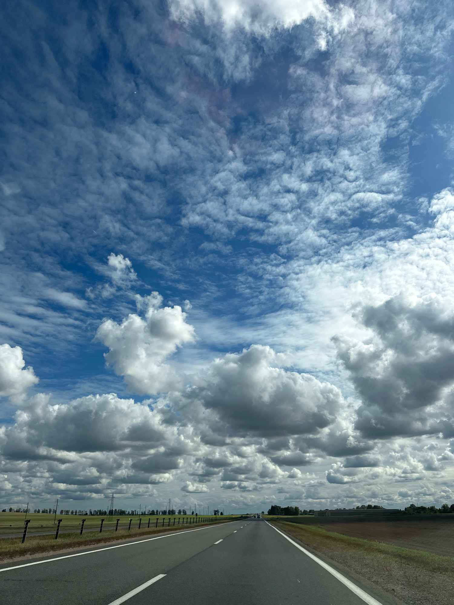 The image depicts a view from a vehicle traveling on a paved road through a rural area. The road has white lane markings. To the left of the road, there is a grassy area with a fence and agricultural fields. To the right, there are brown agricultural fields. In the distance, a line of trees and utility poles are visible. The sky is a prominent feature, filled with a mix of white and gray clouds against a bright blue backdrop. The overall scene suggests a typical day in the countryside. The location is specified as Блонскі сельскі Савет, Belarus.