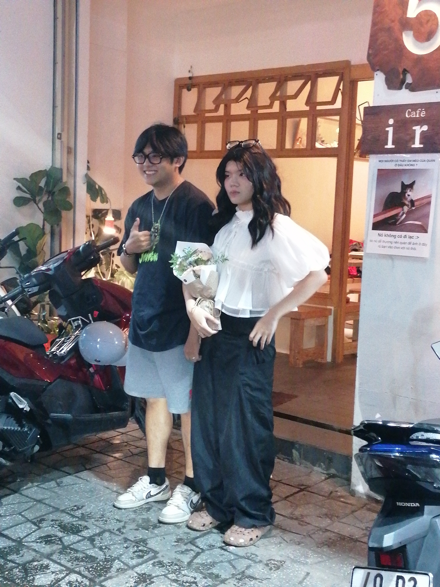 Two young adults, a man and a woman, are standing on a patterned tile sidewalk in front of a building. The man is on the left, wearing glasses, a dark t-shirt, and light grey shorts. He is giving a thumbs-up gesture and holding a bouquet of flowers. The woman is on the right, wearing a white blouse with puff sleeves and a long black skirt. She has long, dark, wavy hair and is looking slightly to her left. To the left of the pair, a portion of a red motorcycle is visible, with a grey helmet resting on its seat. Behind the individuals, the building has wooden architectural elements and a sign with "Café ir" written on it. A poster with an image of a cat and Vietnamese text is also visible on the wall. The environment appears to be an urban setting, likely in Da Lat, Vietnam, given the presence of the cafe sign and the overall aesthetic.