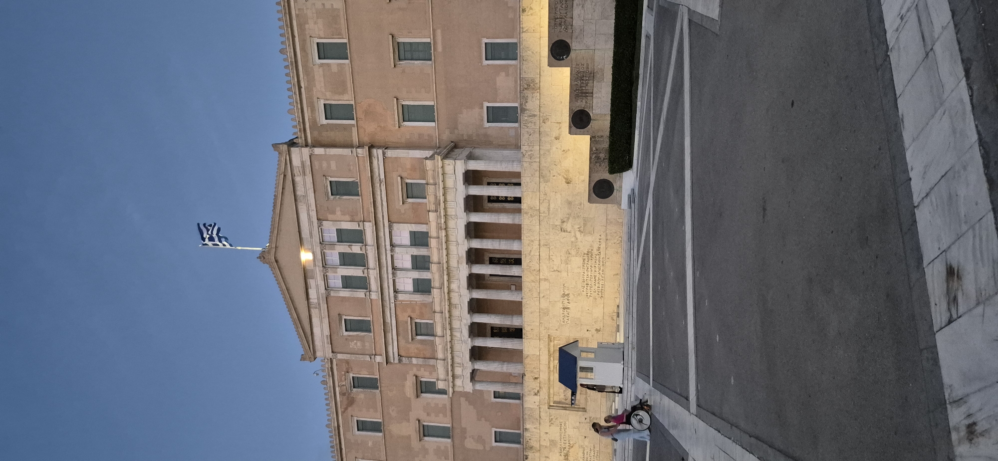 The image shows the Hellenic Parliament building in Syntagma Square, Athens, Greece, under a clear blue sky. The building is a large, Neoclassical structure with a prominent portico supported by columns. A Greek flag is visible flying from a flagpole atop the building. In the foreground, there is a memorial with inscriptions on a stone wall, flanked by circular bronze plaques set into stepped platforms. To the left, a small guardhouse with a blue roof stands near the memorial. Two individuals are present: one is a woman in a light-colored dress standing next to a person in a wheelchair. The ground is paved with asphalt and marble. The scene appears to be captured in the early evening.