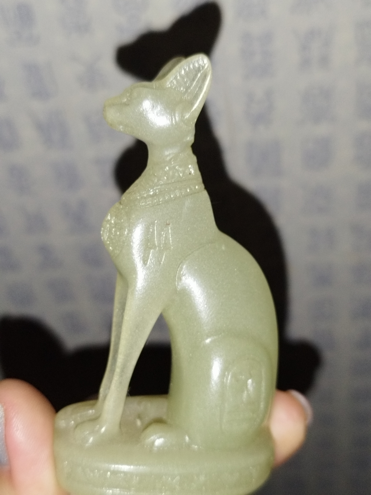 A translucent, pale green figurine of a sitting cat, likely an Egyptian Bastet representation, is held in a person's hand. The figurine has large, pointed ears, a slender neck adorned with a beaded collar, and delicate front legs. The cat sits on a circular base. Visible on the body of the figurine are etched symbols, one resembling hieroglyphs on the chest and another a square emblem on the flank. The figurine casts a prominent shadow on a textured background patterned with faded blue motifs. The lighting suggests an indoor setting, possibly in Saint Petersburg, Russia. The image is a close-up shot, focusing on the detail and texture of the figurine.