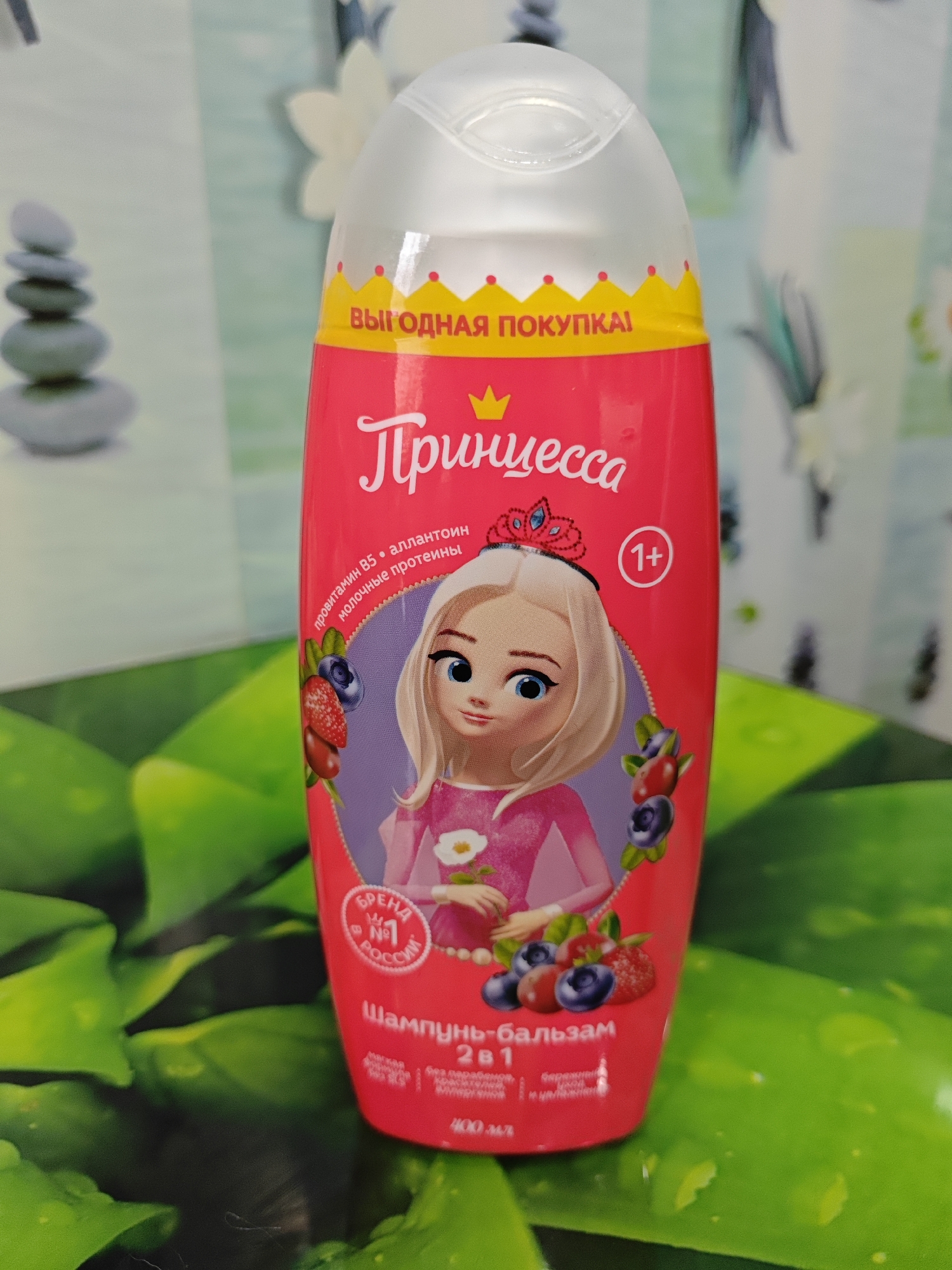 A bright pink bottle of "Princessa" shampoo-balm 2-in-1 is centrally placed on a green, textured surface, possibly a table or mat. The bottle features a cartoon image of a blonde princess with blue eyes, wearing a pink dress and a tiara, surrounded by berries. The text on the bottle is in Russian and indicates it is for ages 1+. The background is blurred and features a light-colored pattern with floral motifs on the left and a darker, abstract pattern on the right. There is no indication of the city of Polyarny, Russia, in the image or its context.