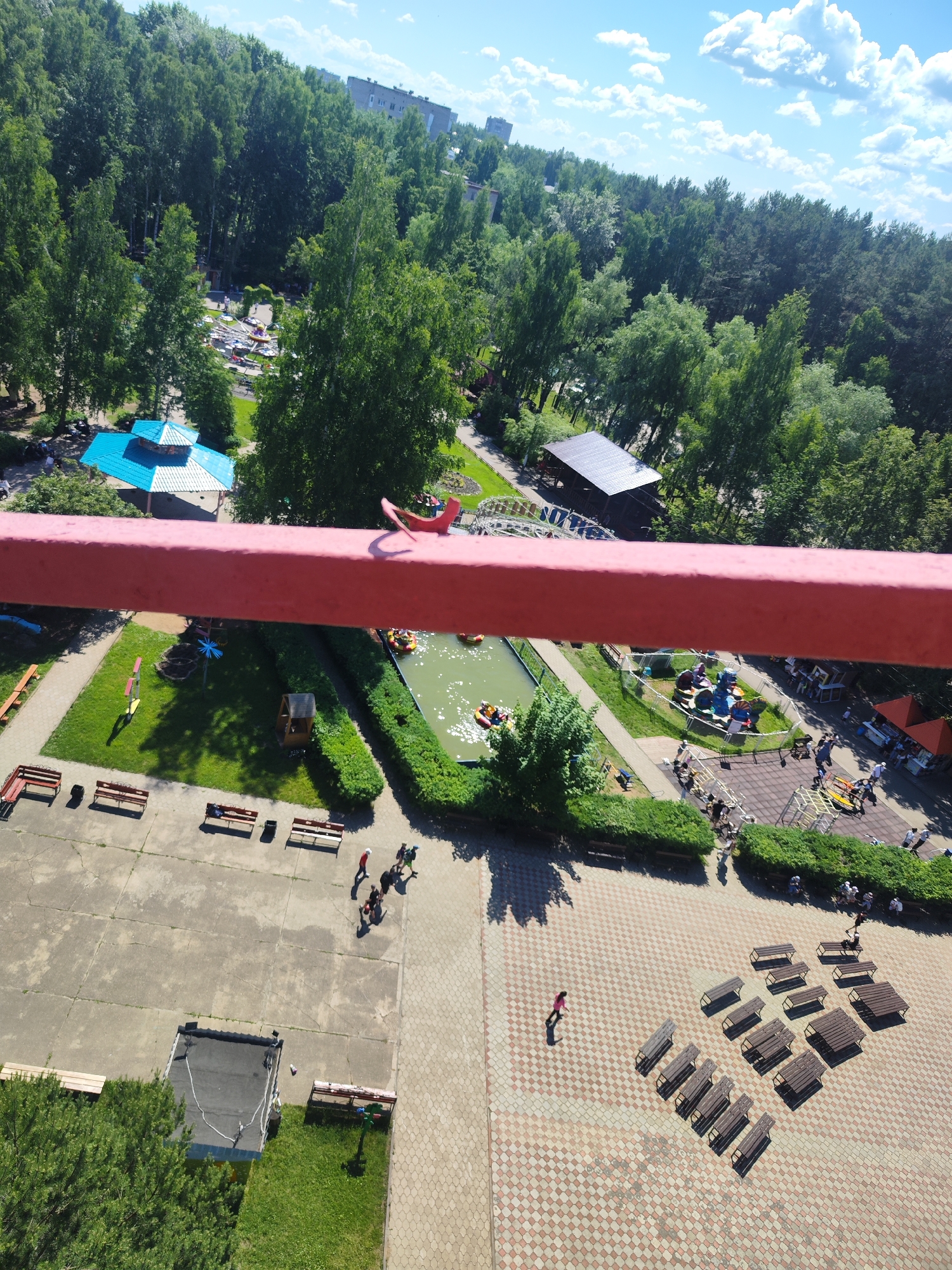 This aerial photograph, taken in the city of Chaikovsky, Russia, captures a vibrant amusement park on a clear, sunny day. The foreground is dominated by a large, red, horizontal beam, likely part of a Ferris wheel or similar attraction, obscuring the lower portion of the image. Below this beam, the park unfolds with a variety of attractions and amenities.
To the left, a bright blue and white gazebo with a tiered roof stands amidst lush green trees. Adjacent to it is a water park area with several yellow inflatable boats floating on a body of water. Further to the right, a small playground area features colorful rides, and beyond that, a picnic area with many wooden tables and benches is visible. People are scattered throughout the park, engaged in various activities, including walking, riding the attractions, and sitting at the picnic tables.
The background is a dense expanse of green trees, indicative of a wooded park or forest. In the distance, buildings can be seen peeking through the trees, suggesting the park's location within a populated area of Chaikovsky. The sky is a clear blue, dotted with fluffy white clouds, contributing to the overall bright and cheerful atmosphere of the scene. The ground surfaces vary from paved areas to grassy lawns and the patterned tiles of the picnic area.