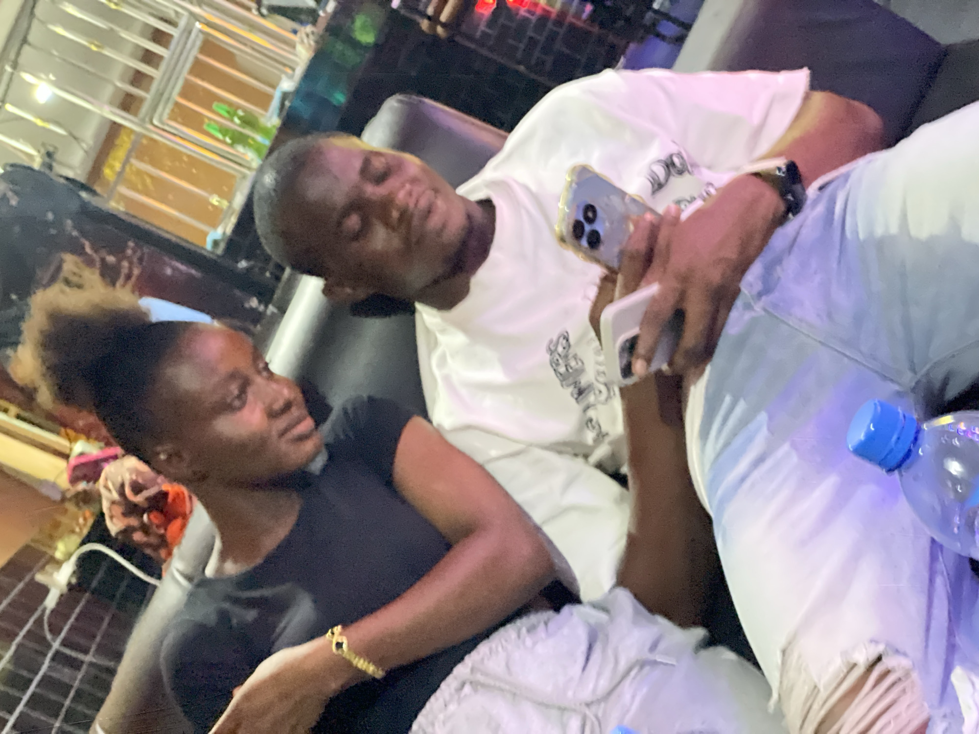 In a dimly lit environment, likely in Obosi, Nigeria, a woman with dark hair and a black top sits on a dark leather couch, looking towards a man reclining beside her. The man, wearing a white shirt, holds two smartphones in his hands and appears to be looking at one of them. He also wears a dark wristwatch on his left wrist. A water bottle with a blue cap is visible near his ripped light-colored jeans. Behind them, a reflective structure with visible objects inside can be seen.