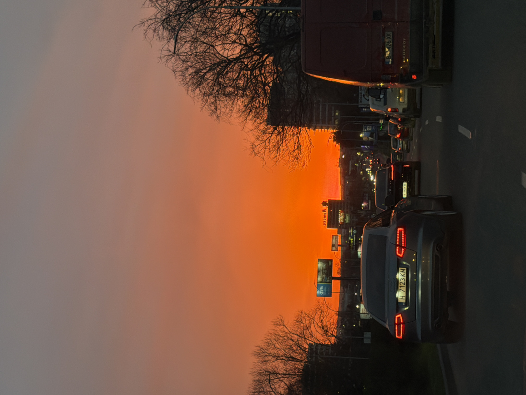 The image shows a street in Varna, Bulgaria at dusk. An Audi car with the license plate H 7123 KM is prominently featured, positioned on the right side of the road. A line of traffic extends ahead, including a red Peugeot van. Bare trees flank the street, silhouetted against a vibrant orange sunset. Buildings and street signs are visible in the distance, illuminated by the setting sun.
