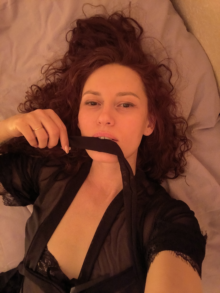A female subject with reddish-brown, curly hair lies supine, looking directly into the camera. She wears a sheer black robe with lace trim on the sleeves, partially revealing a black lace bra. Her left hand, bearing a thin gold-colored ring on the ring finger, holds a black fabric strip, which is partially inserted into her slightly open mouth. The background consists of a light-colored fabric, consistent with bedding. The scene takes place in an indoor setting, in the city of Saint Petersburg, Russia.