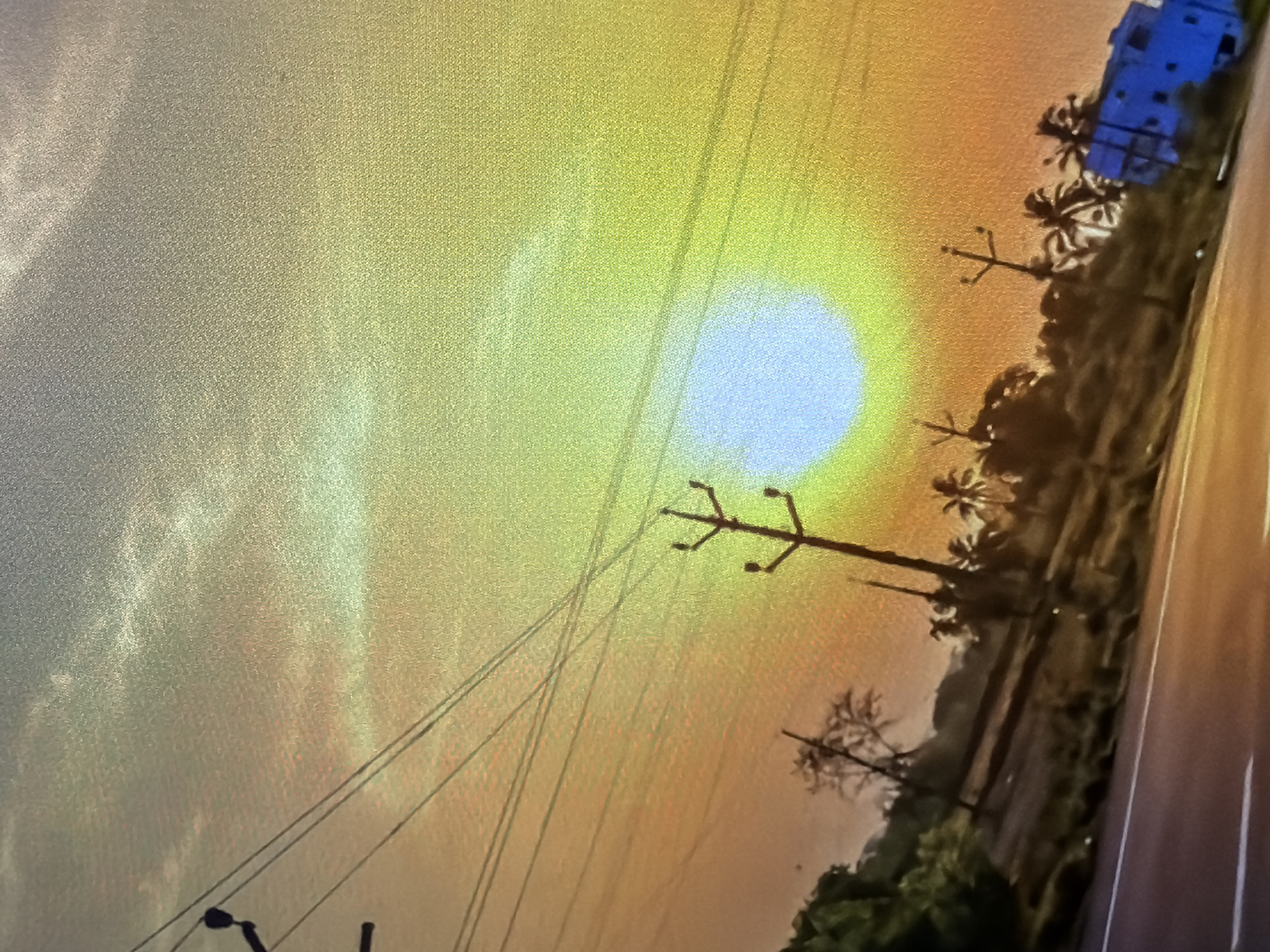 The image depicts a sunset over a rural or semi-rural landscape. A bright, hazy sun, appearing almost white, is central in the sky, surrounded by a gradient of warm colors ranging from yellow and green at the top to orange and red towards the bottom. Several utility poles with wires are visible, running diagonally across the frame, suggesting the presence of a road or a populated area nearby. In the middle ground, silhouetted trees, including palm trees, are scattered across a slightly elevated terrain. To the right of the frame, a blue, boxy structure, possibly a building or a vehicle, is partially visible. The foreground shows a dark surface, likely a road, with a white line marking its edge. The overall atmosphere is hazy and warm, typical of a sunset.