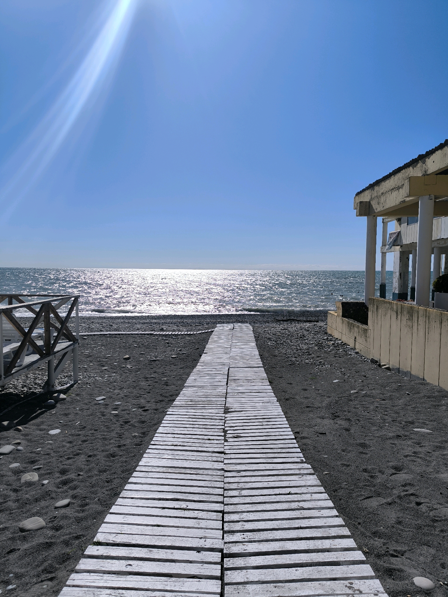 A bright, sunny day on a black sand beach in Sochi, Russia. A white wooden boardwalk extends from the foreground towards the sparkling, sun-drenched sea. On the right, a building with a beige facade and white columns is partially visible. To the left, a white wooden railing frames the beach. The sky is a clear, vibrant blue, with a sunbeam cutting across the upper left corner. The sea is calm, with small waves lapping at the shore. The beach is made up of dark sand and smooth pebbles.