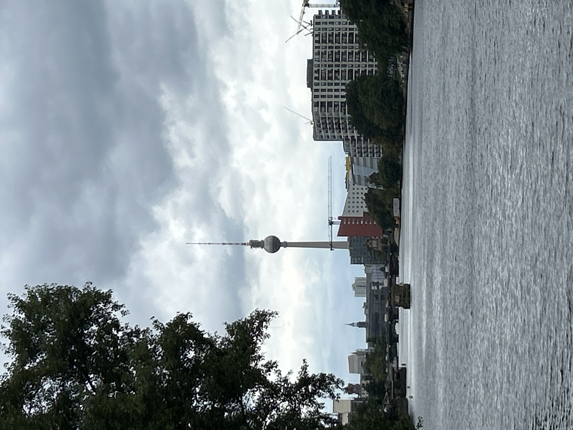 The image shows a body of water, likely a river, in the foreground. In the middle ground, there is a cityscape with various buildings, including a tall, slender tower with a spherical section and antenna at the top, characteristic of the Fernsehturm (TV Tower) in Berlin, Germany. A bridge can be seen spanning the river. Further back, more buildings and trees are visible. The sky is overcast with gray clouds. The scene appears to be an urban landscape along a river, with the Fernsehturm serving as a prominent landmark. There are no people or discernible actions captured in the image. The overall atmosphere is somewhat muted due to the cloudy sky.