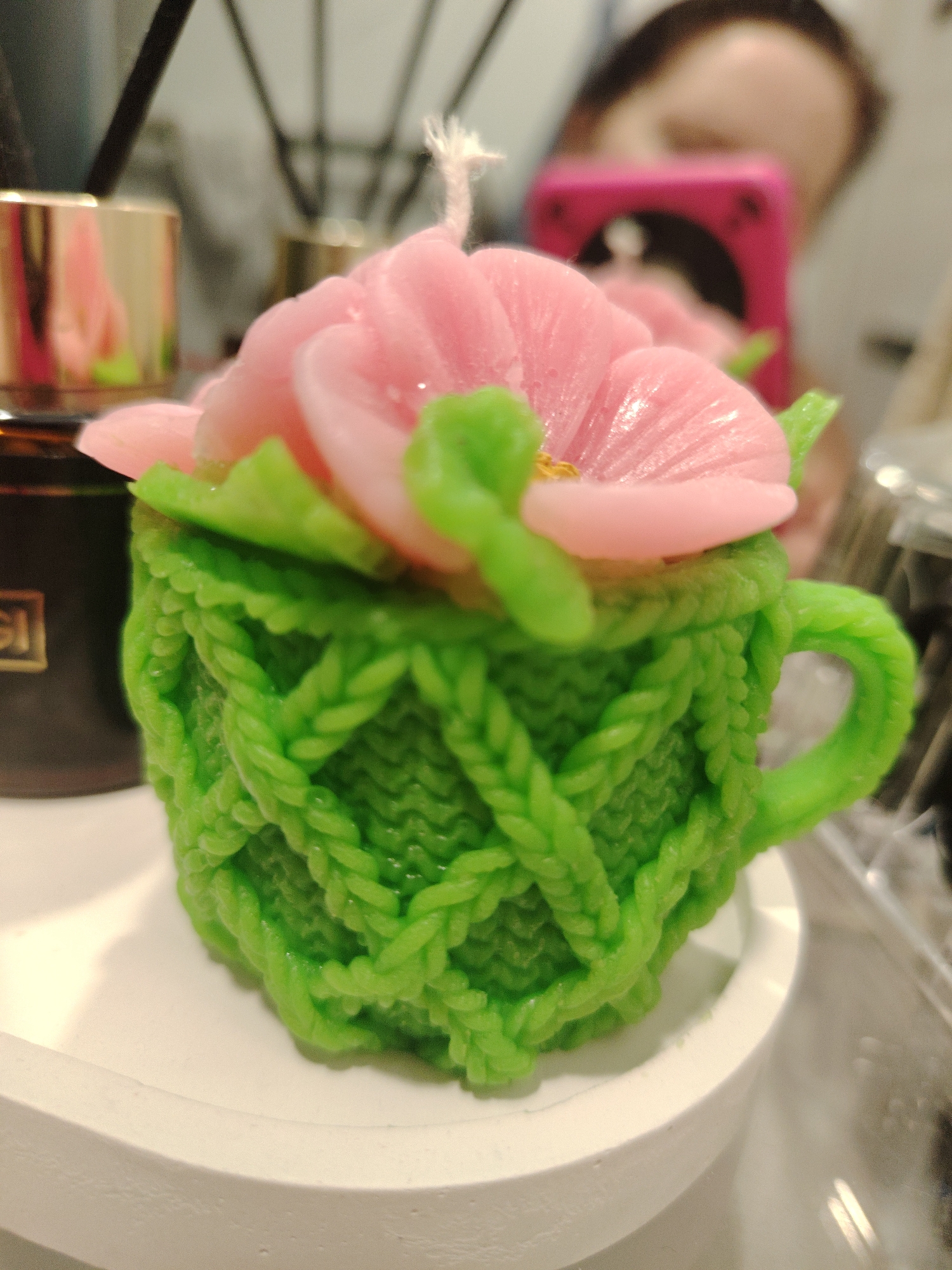 A close-up photograph captures a handcrafted candle resembling a tea cup. The candle is bright green, textured to mimic knitted yarn. Pink flower-like petals adorn the top, with green leaves nestled beneath them. The candle sits on a small, white circular tray. In the background, out of focus, are a brown bottle with a gold-colored cap, what appears to be a diffuser, and a person partially visible using a pink mobile phone. The scene is well-lit, and the details are sharp, suggesting a modern, indoor setting. There is no indication of the location Sarapul, Russia.